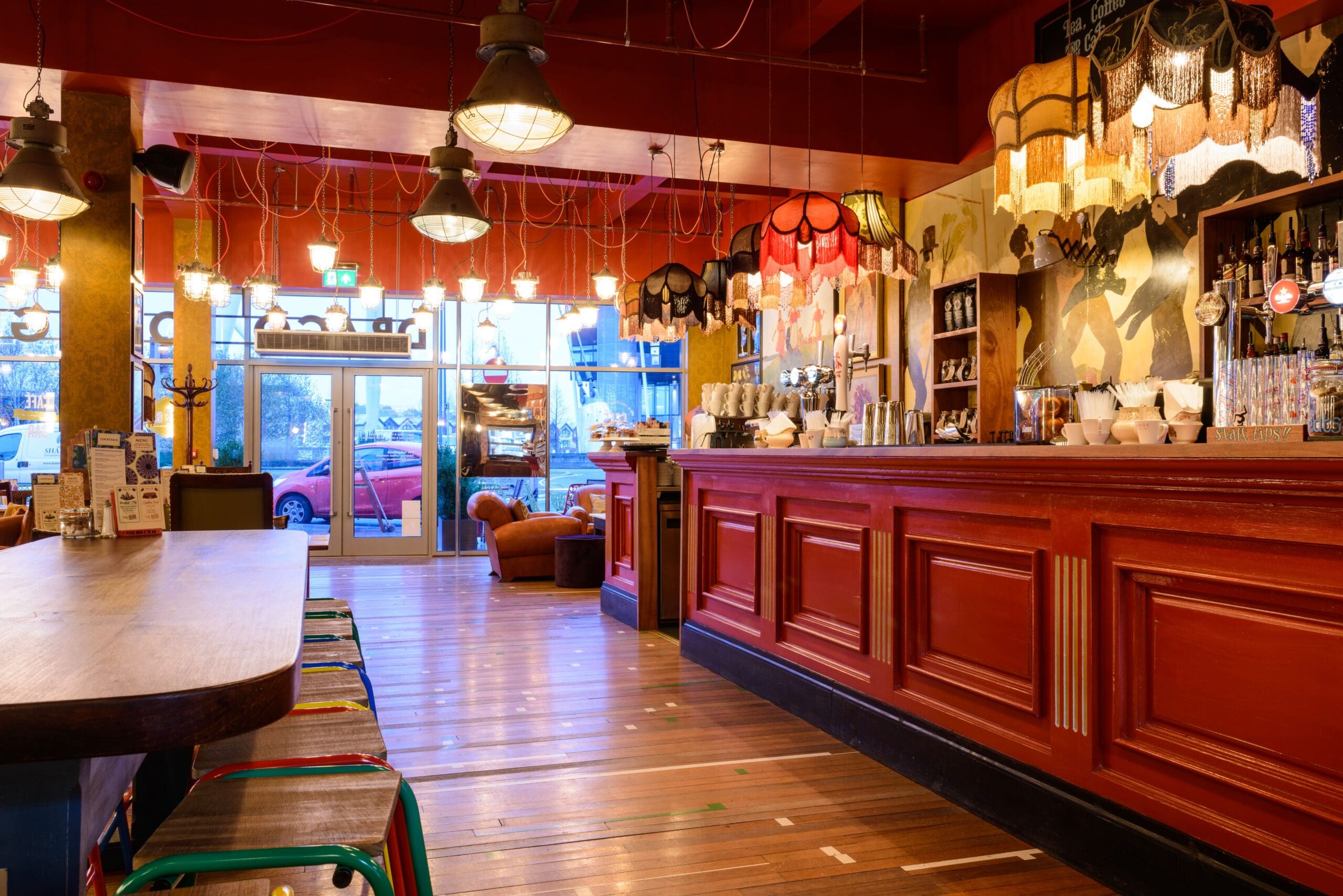 A cozy, warmly lit bar named Drago features red wooden panels, colorful hanging lamps, a long bar counter, shelves with bottles, high stools, and large windows overlooking parked cars outside.