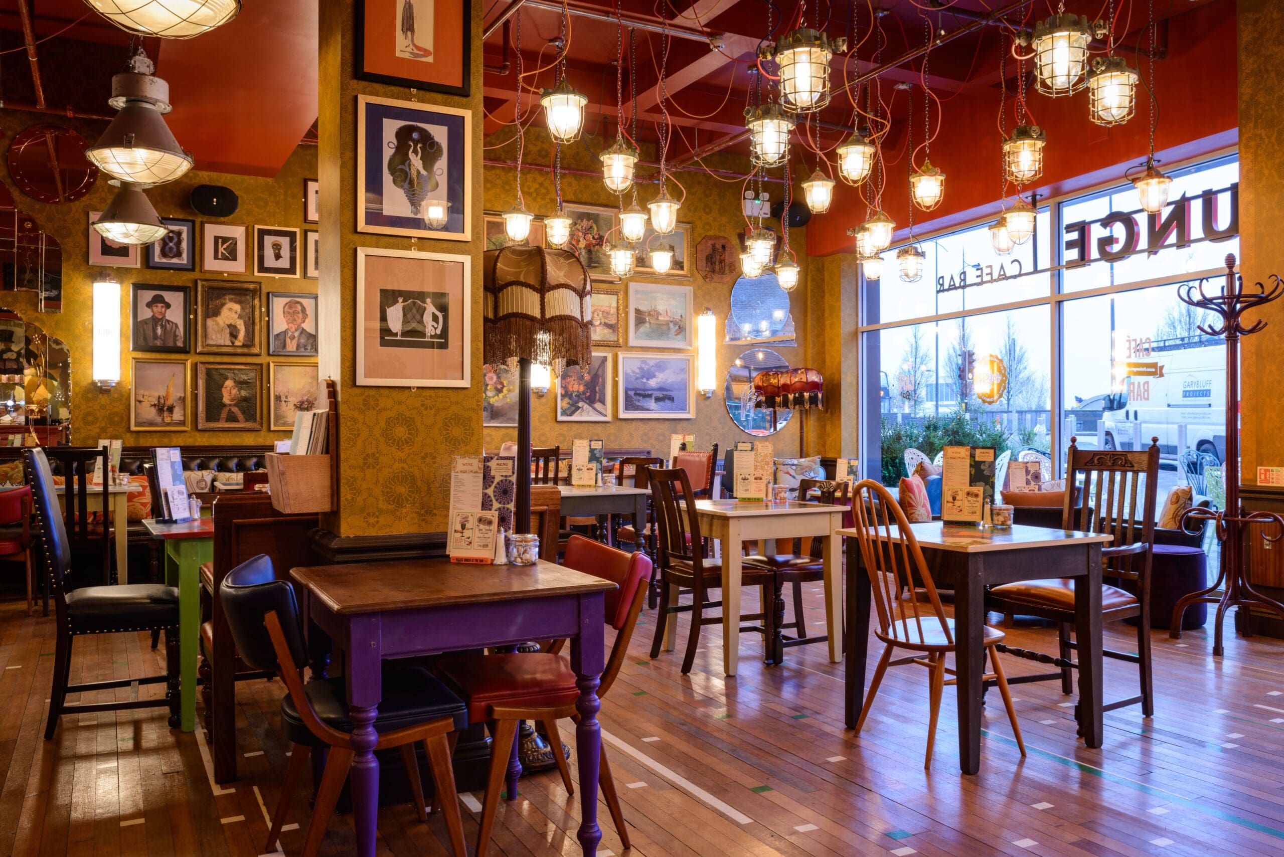 A cozy, eclectic café interior with mismatched wooden tables and chairs, warm lighting from hanging lamps, Drago-inspired framed art on yellow patterned walls, and large windows letting in natural light.