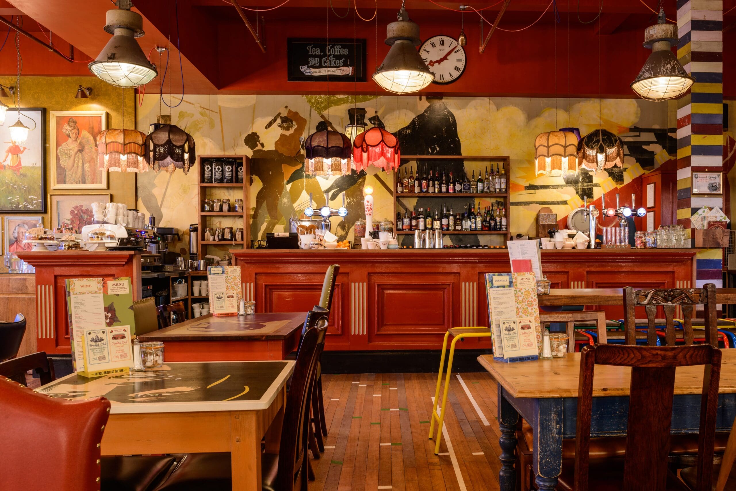 Cozy café interior with colorful lamps, eclectic Drago decor, wooden tables and chairs, a red counter, shelves of bottles and mugs, and a large wall mural behind the bar. Warm lighting creates a welcoming atmosphere.