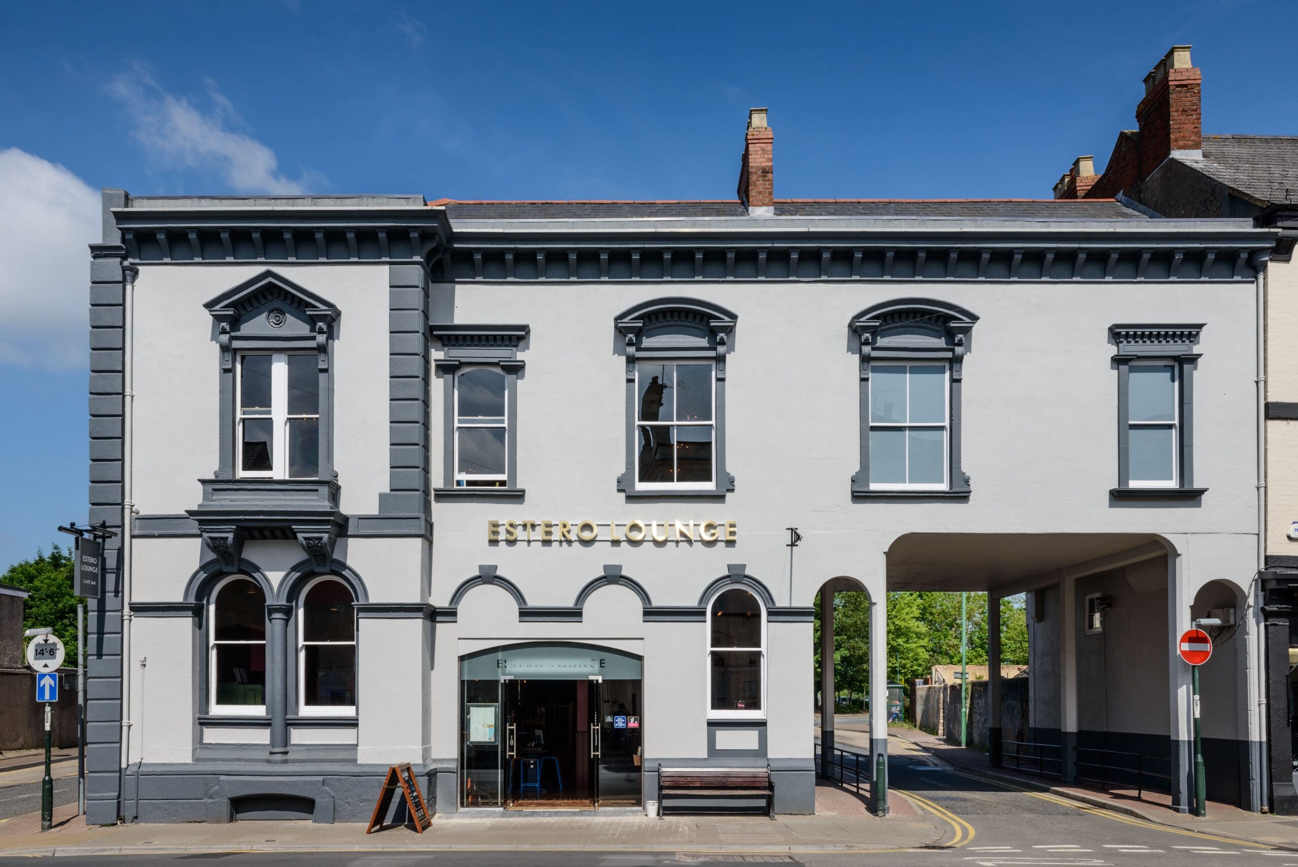 A two-story, light gray building with arched windows and decorative trim houses Bistro Lounge in Estero. An archway on the right leads to a side street. The sky is clear and blue.