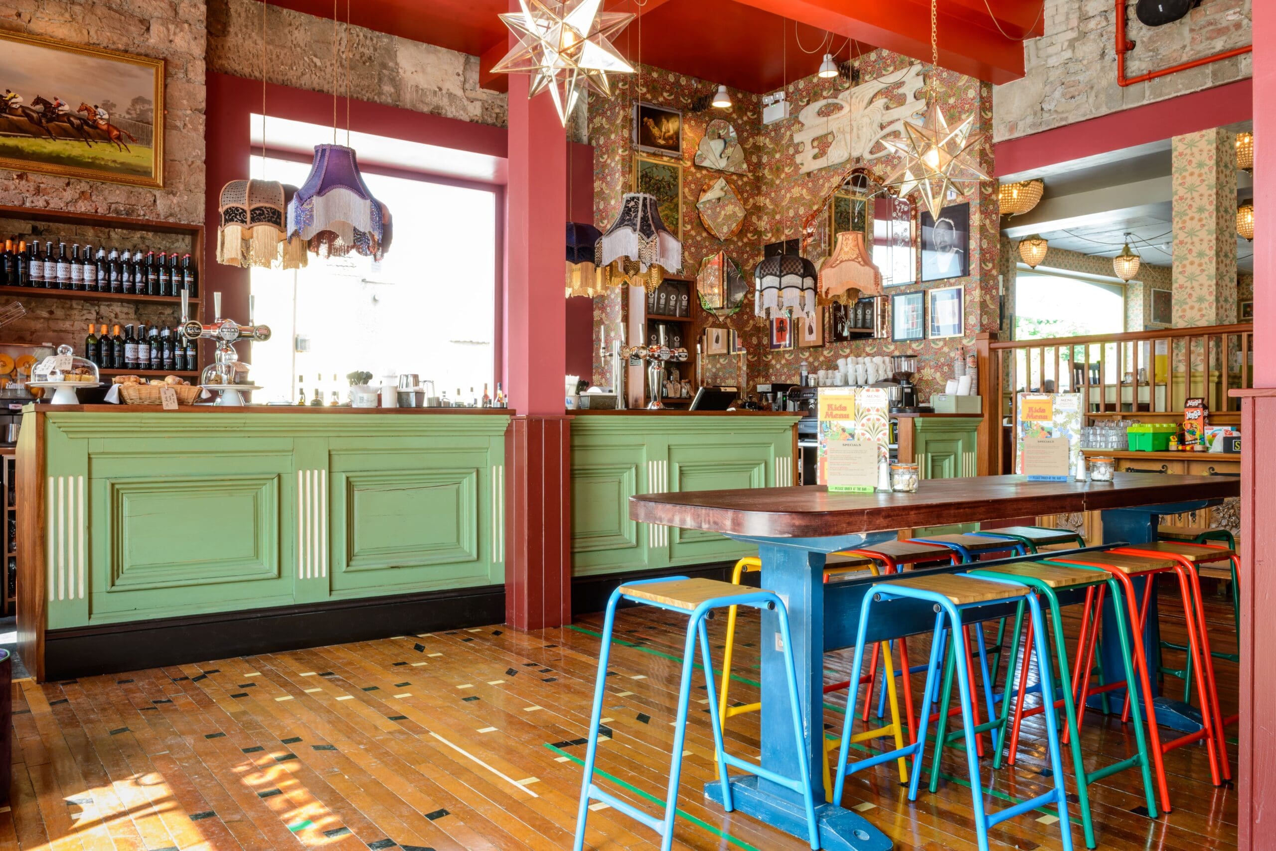 Brightly lit Estero cafe with a green bar counter, colorful bar stools, wooden floor, eclectic hanging lamps, star-shaped light fixtures, and vintage decor. Shelves with bottles and art adorn the exposed brick walls.
