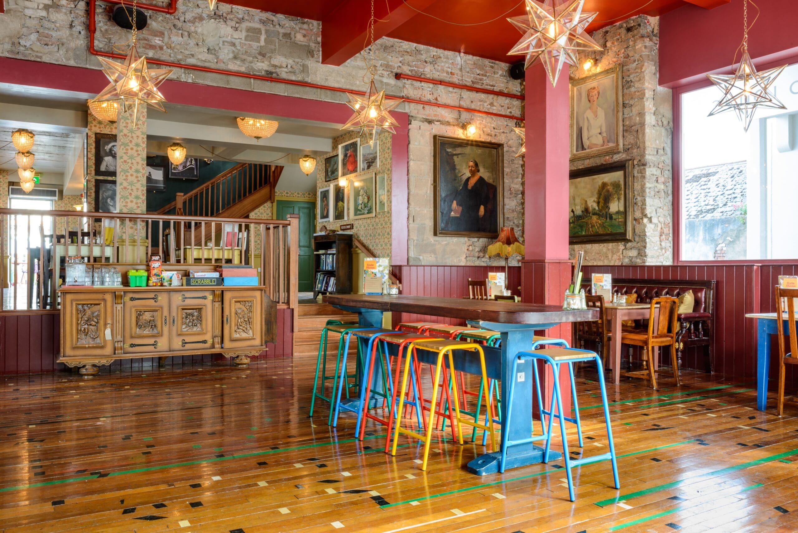 Colorful bar stools surround a long table in a bright, eclectic Estero café with star-shaped pendant lights, wood floors, brick walls, vintage art, and large windows letting in natural light.