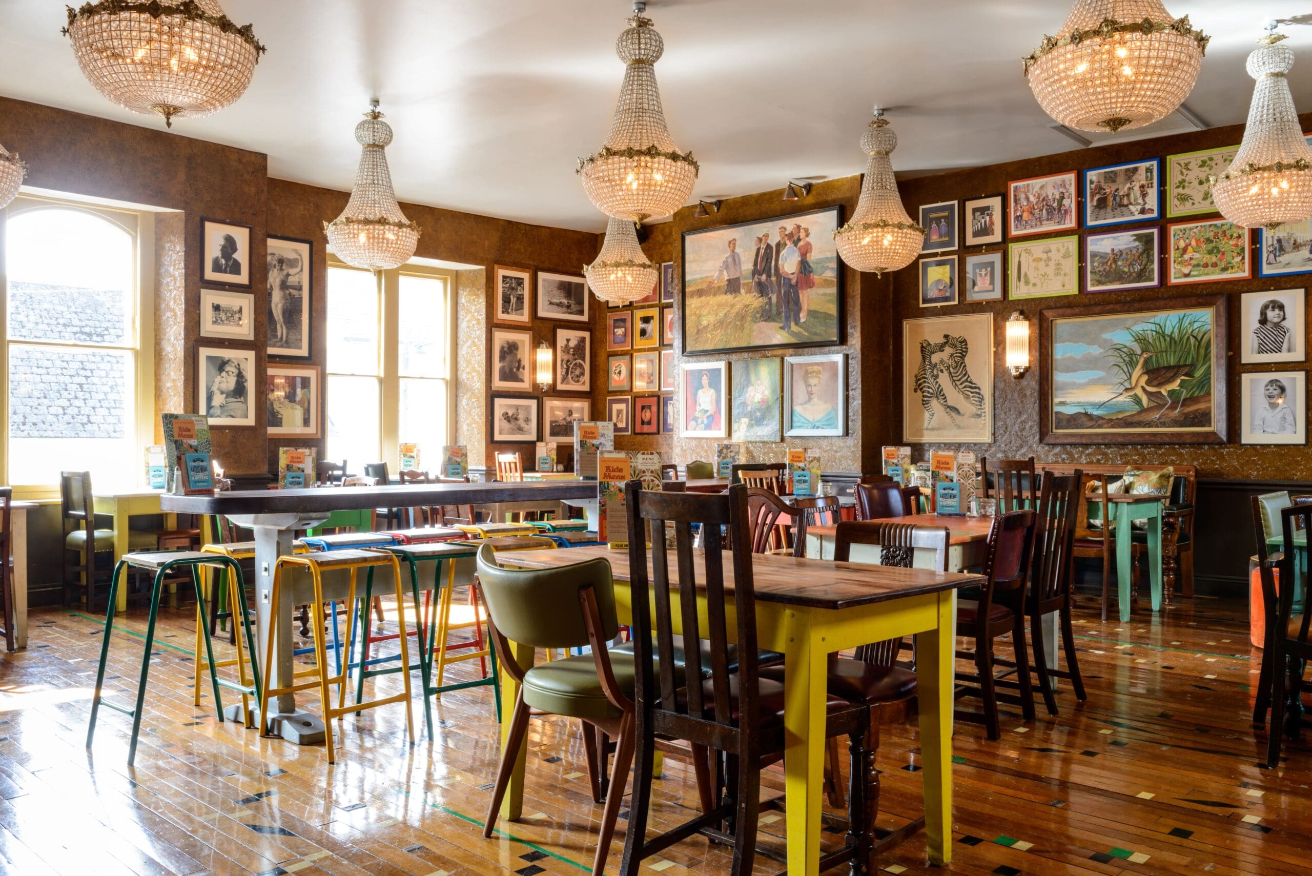 Bright, eclectic restaurant interior in Estero with mismatched wooden chairs and tables, chandeliers, large windows, and walls covered in various paintings and framed art in different styles and sizes. Polished wood floor.