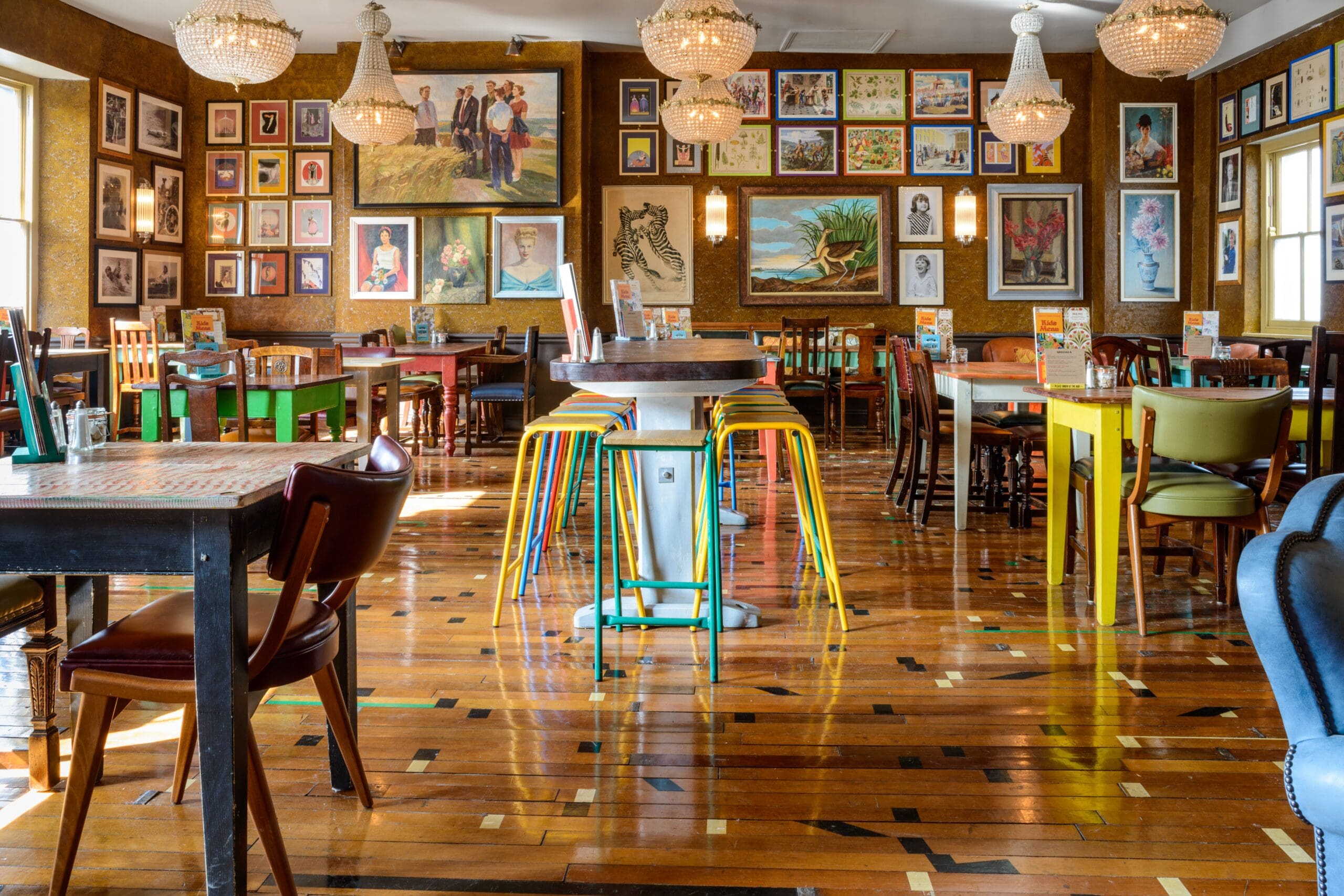 A colorful, eclectic restaurant interior in Estero features mismatched chairs and tables, wooden floors, and walls filled with framed art and paintings. Crystal chandeliers hang from the ceiling, creating a cozy, artistic atmosphere.