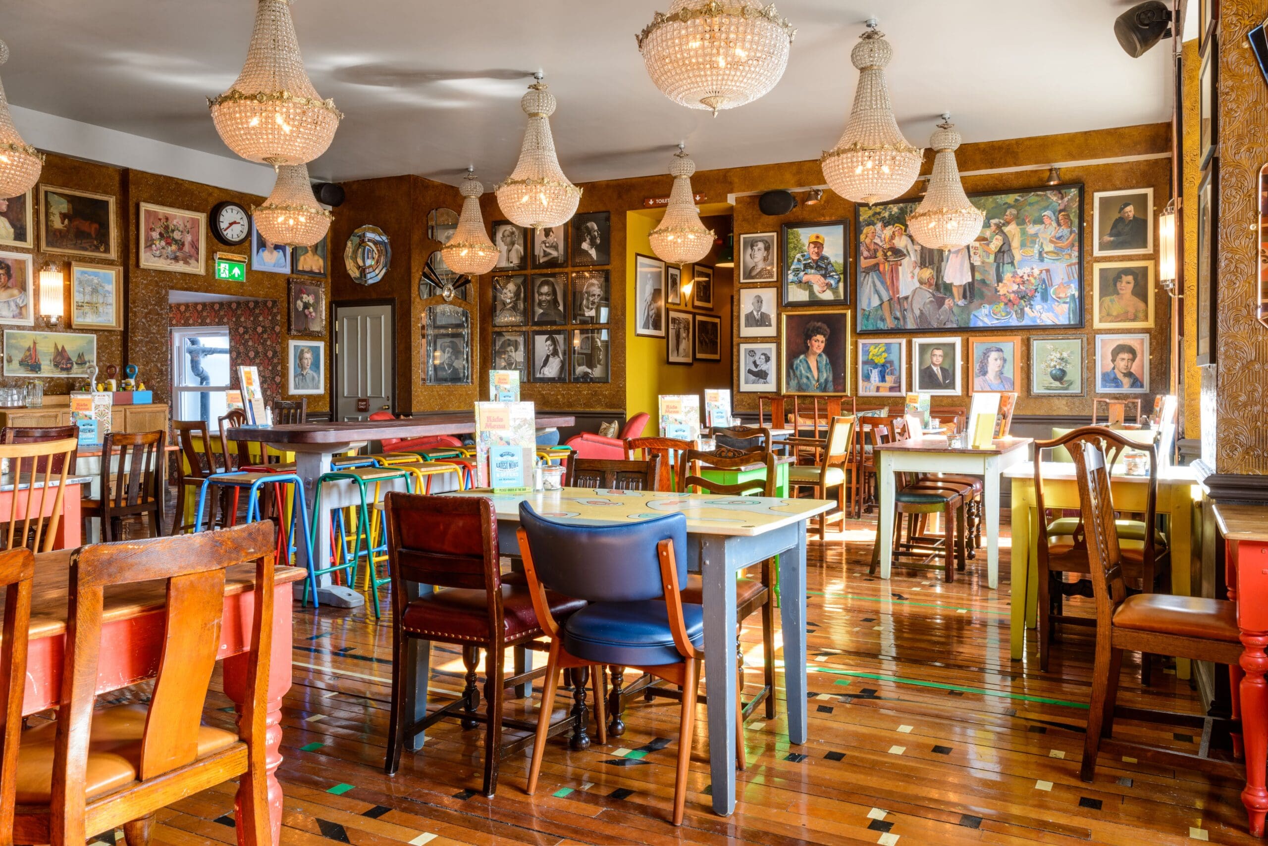 A bright, eclectic restaurant interior in Estero with colorful chairs, wooden tables, chandeliers, and walls covered in framed portraits and artwork. Sunlight streams through the windows, creating a warm and inviting atmosphere.