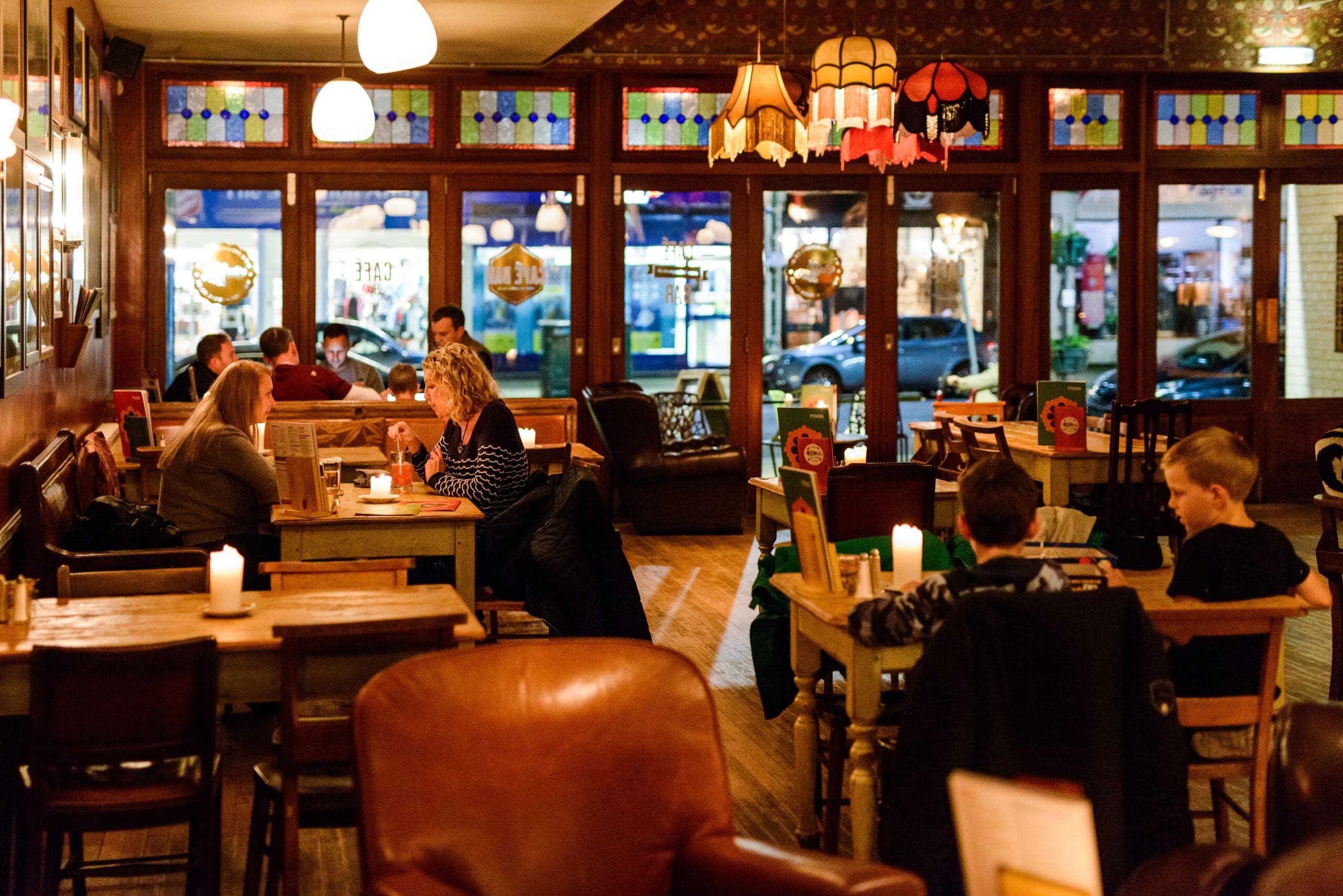 Cozy restaurant interior with wooden tables, stained glass windows, and warm lighting. Several people dine and converse while playing Ludo. Candles on tables add to the relaxed, inviting atmosphere.