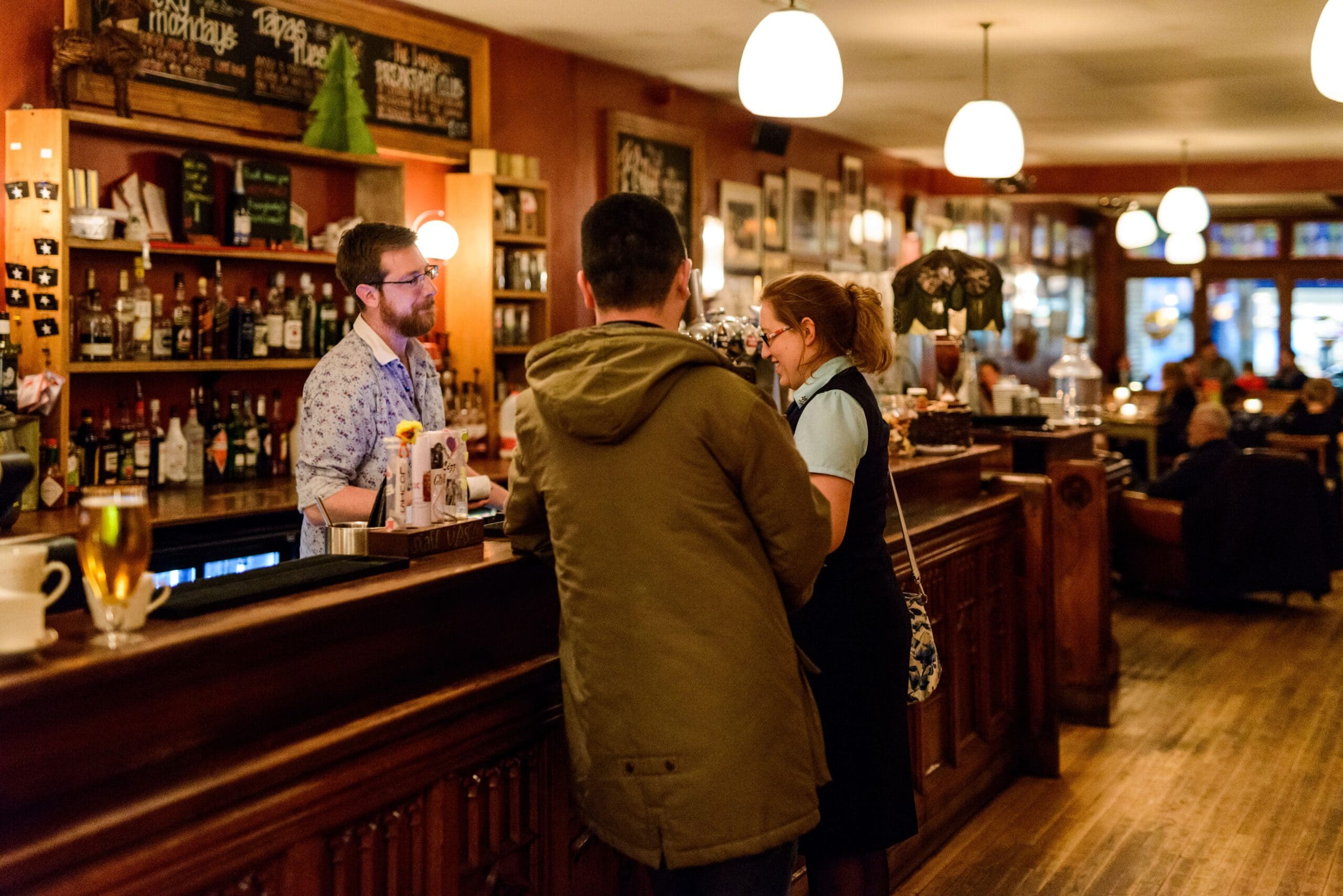 A bartender chats with two customers at a wooden bar in a warmly lit, busy restaurant or pub, where cozy decor and Ludo boards add to the charm. Other patrons are seated at tables in the background, enjoying the inviting atmosphere.