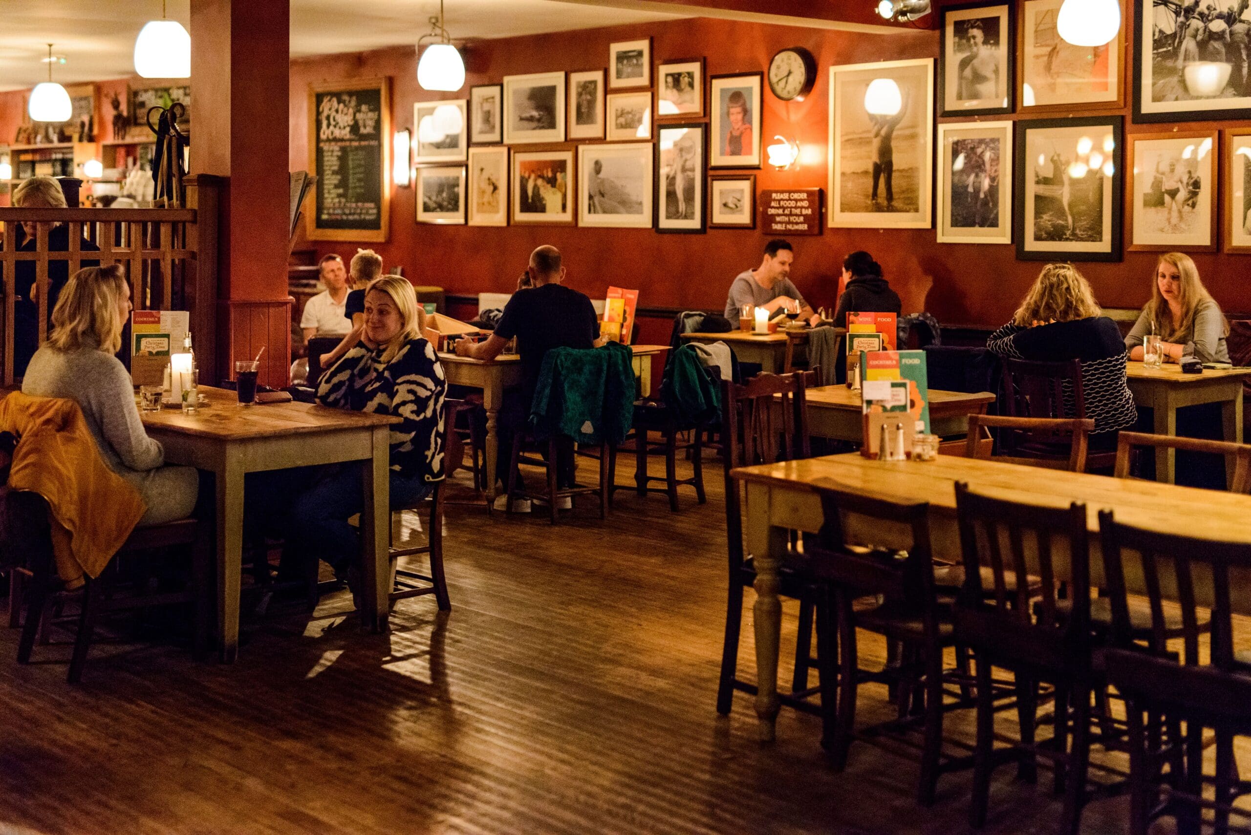 A cozy restaurant with wooden tables and chairs, warmly lit by hanging lights. People are dining, chatting, and playing Ludo in small groups. Framed photos and art decorate the reddish-brown walls. The atmosphere is relaxed and inviting.