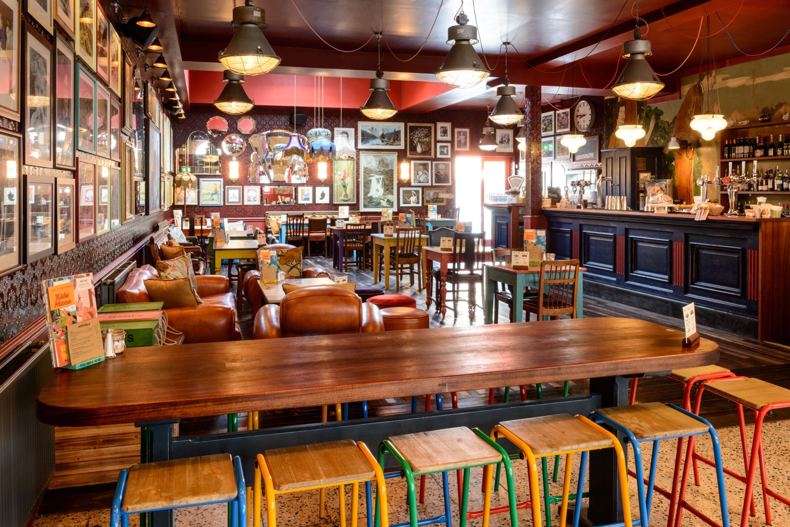 A cozy, eclectic pub with a long wooden table, colorful stools, leather armchairs, framed art on the walls, and warm pendant lighting above the bar and dining area—a true Nautico gem.