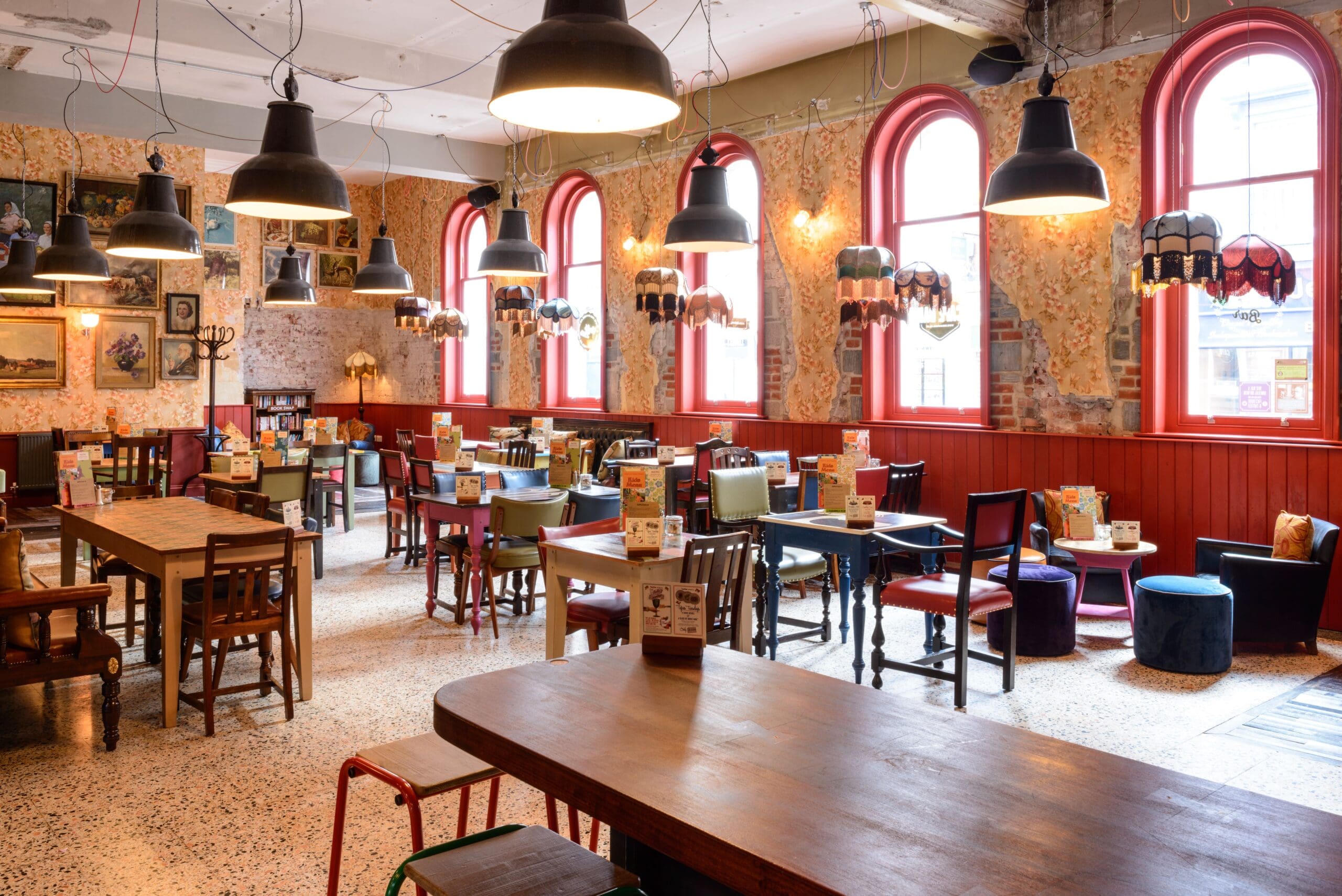 A cozy, eclectic cafe with colorful chairs and wooden tables, vintage decor on patterned walls, large arched windows with red frames, and hanging pendant lights creates a Nautico vibe in this bright, inviting space.