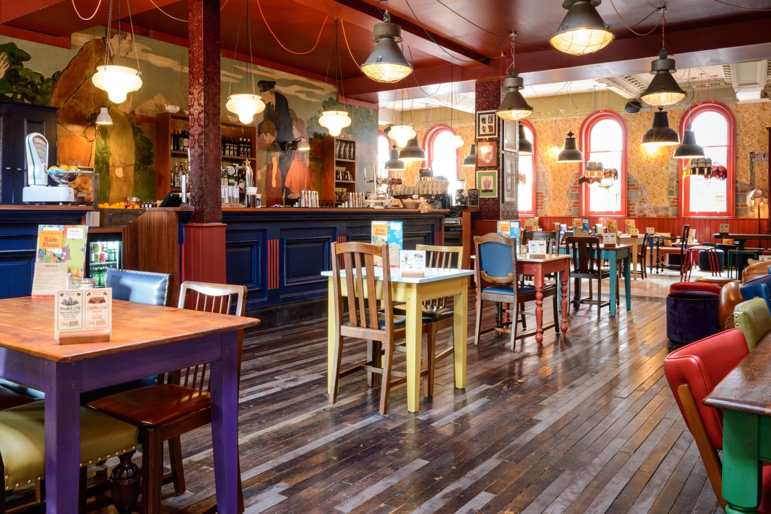 A colorful, eclectic pub interior with mismatched chairs and tables, wooden floors, vibrant Nautico-inspired walls, hanging lights, and a bar area in the background. Sunlight streams through large arched windows.