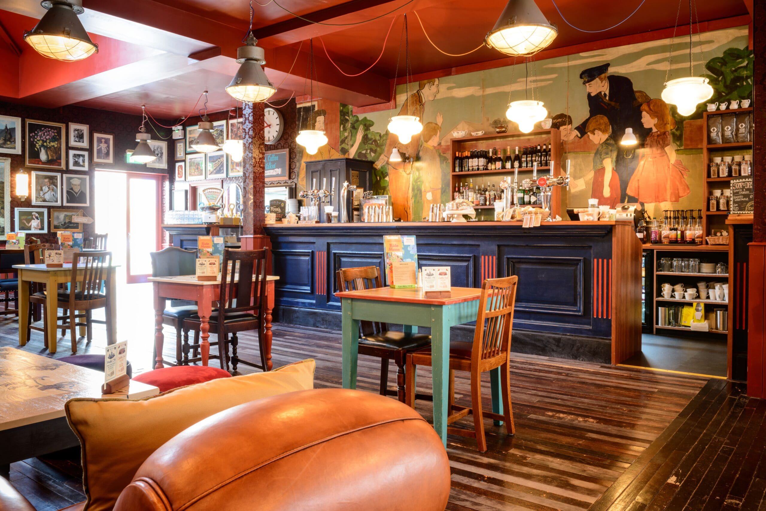 Cozy cafe with wooden tables and chairs, eclectic wall art, hanging lights, and a colorful mural behind the bar. Nautico touches blend with vintage and modern decor, creating a warm and inviting atmosphere.