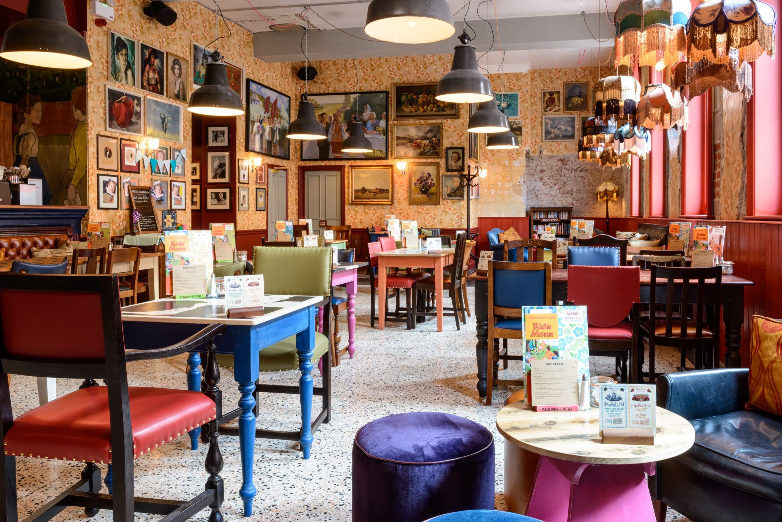 A cozy, colorful café with mismatched chairs and tables, eclectic wall art, Nautico accents, hanging lamps, and menus on each table. Sunlight streams through large windows, creating a warm and inviting atmosphere.