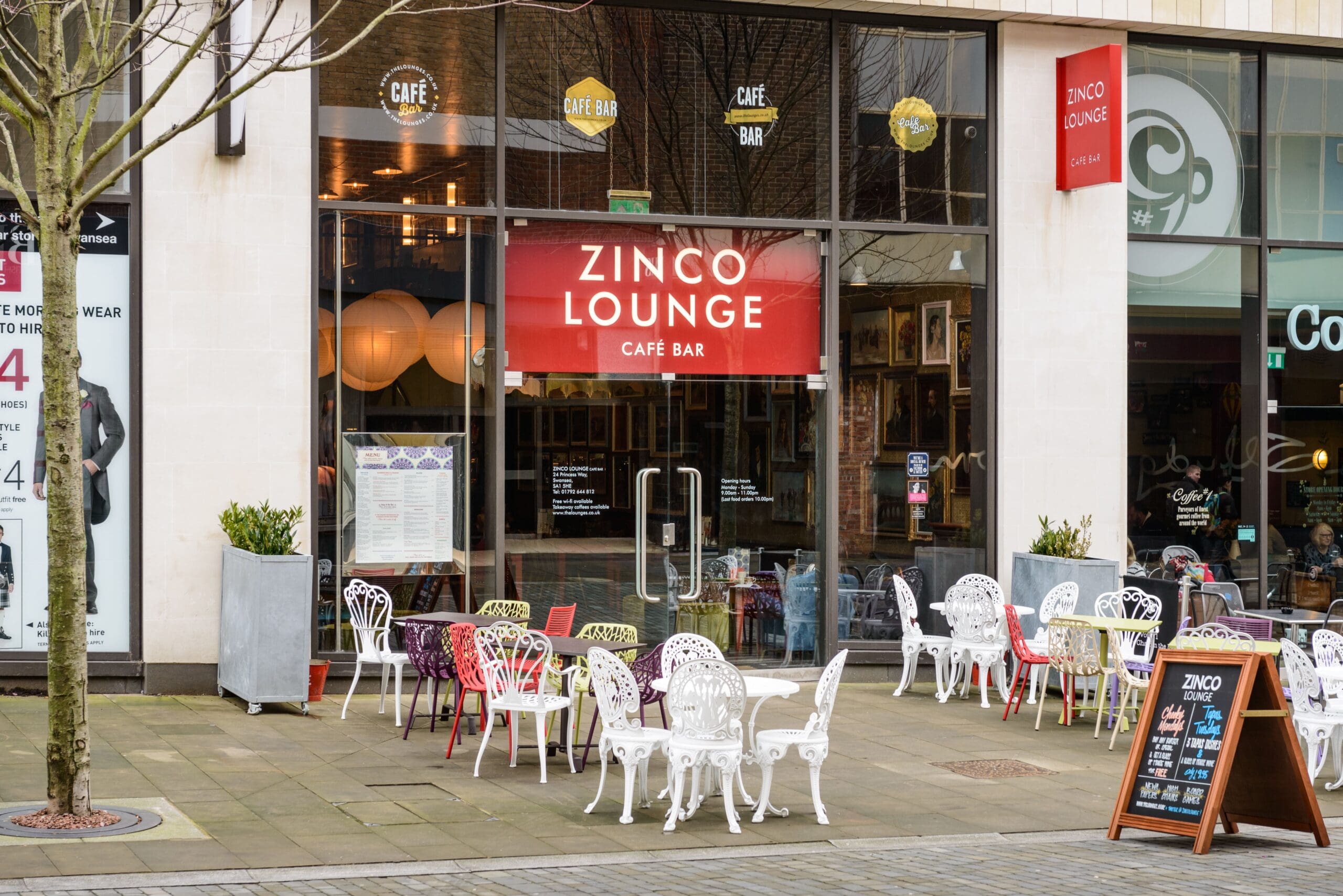 The exterior of Zinco Lounge café bar, with glass doors and windows, stylish Zinco branding, outdoor seating of white and pink metal chairs and tables, and a red sign above the entrance. A menu board and tree are visible on the pavement.