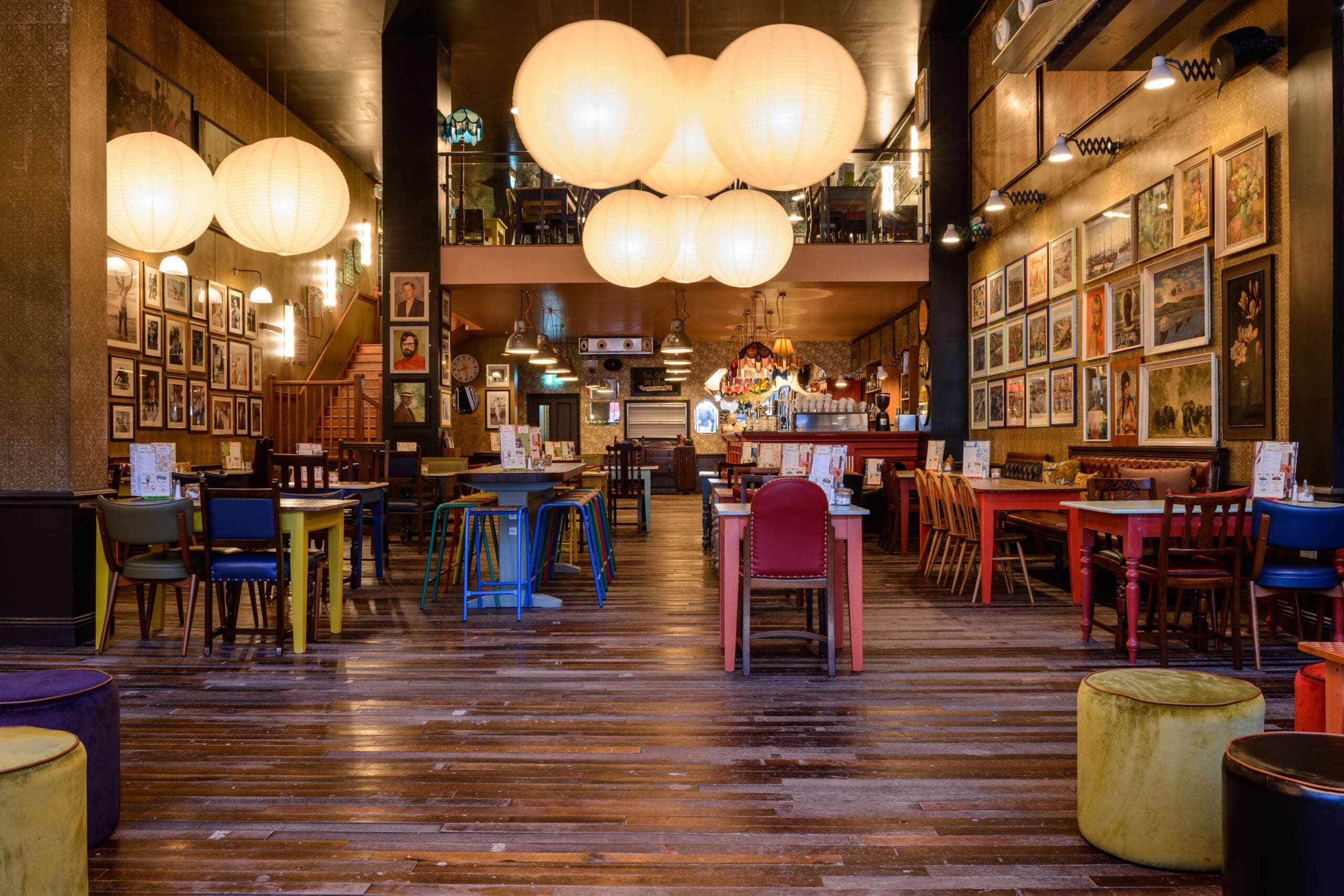A cozy, eclectic restaurant interior with large round pendant lights, colorful mismatched chairs and tables, warm wooden floors, and walls decorated with numerous framed photos and artwork creates the inviting charm of Zinco.