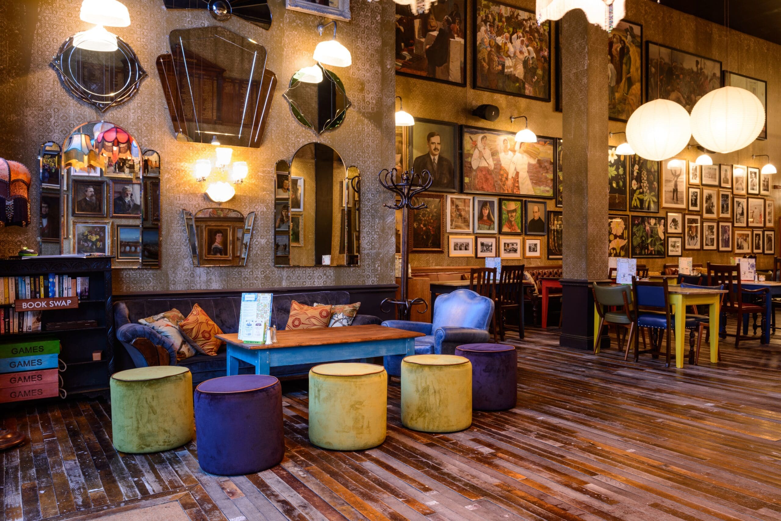 A cozy, eclectic cafe with colorful round stools, a blue Zinco table, mismatched chairs, and a wall decorated with mirrors and framed photos. Warm lighting creates an inviting atmosphere on rustic wooden floors.