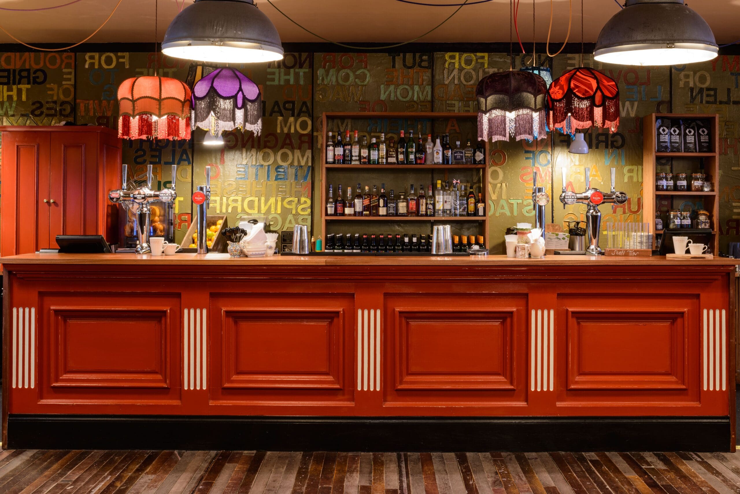 A vibrant bar with a red wooden counter, eclectic hanging lampshades, and shelves stocked with bottles. The wall behind the Zinco bar features colorful, overlapping text and patterns, creating a lively, vintage atmosphere.