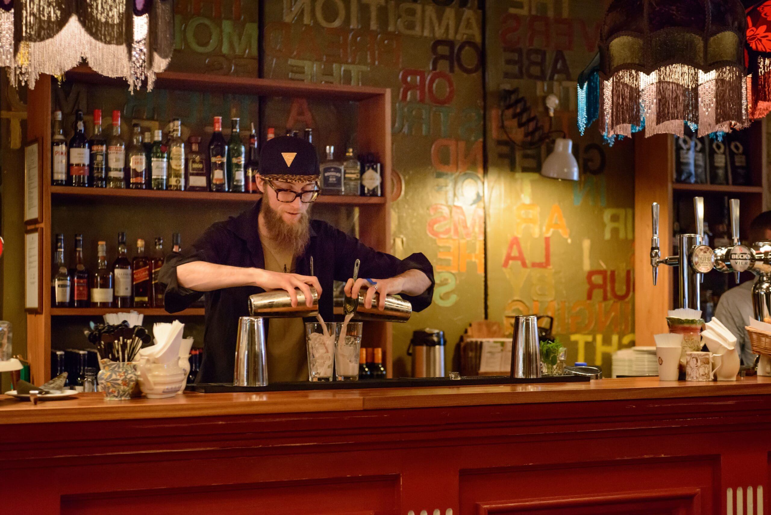A bartender with a long beard and black hat pours drinks from two shakers into a glass behind the vibrant Zinco bar, lined with bottles, glasses, and colorful decor under hanging lamps.