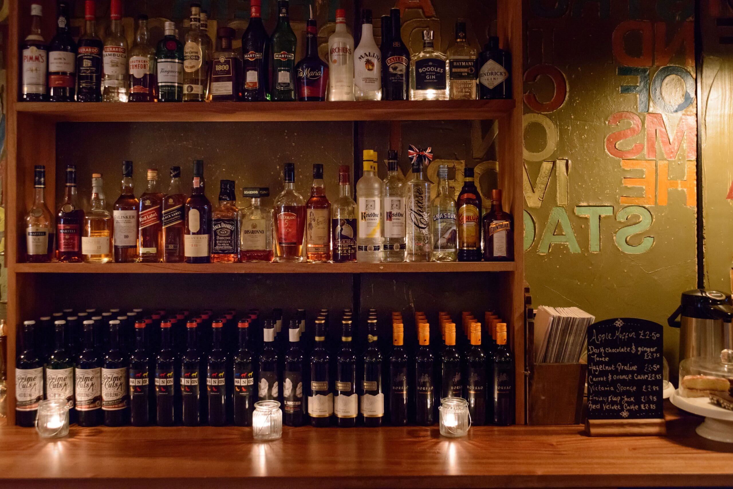 A well-lit bar shelf displays bottles of liquor, rows of Zinco wine, and candles on a wooden counter. A chalkboard menu and glass coffee pot are visible on the right side.