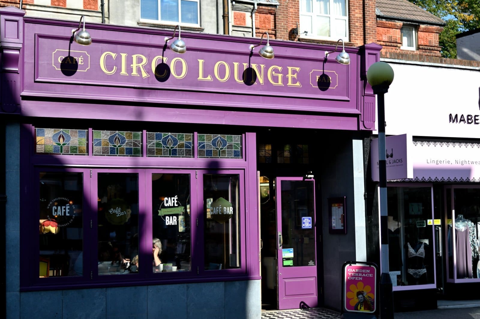 The exterior of Circo Lounge café, painted purple with gold lettering, stained glass windows, and a glass door. Next door is a lingerie and nightwear shop. Sunlight casts soft shadows across Circo’s inviting storefront.