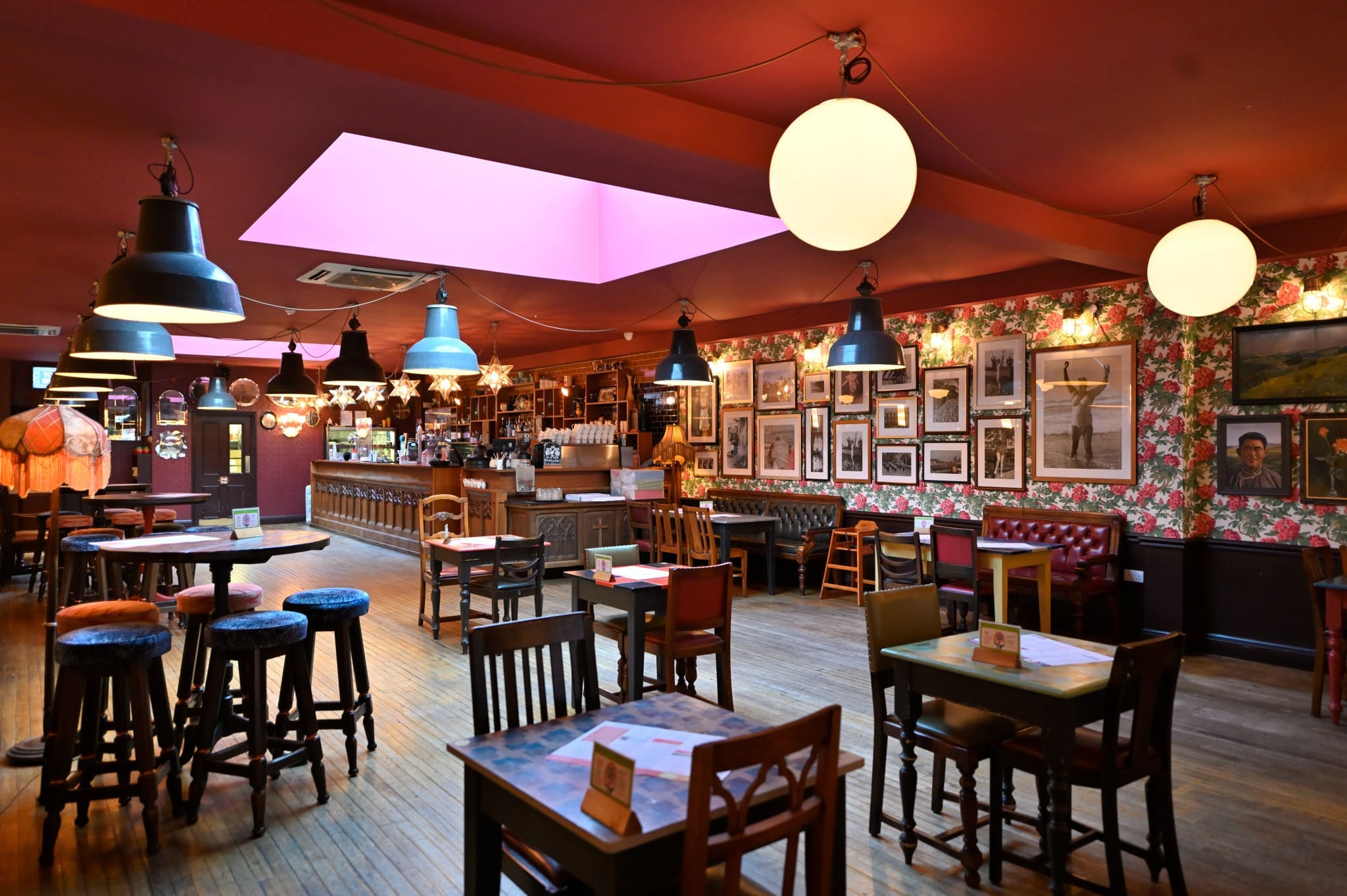 A cozy pub with wooden tables and chairs, colorful wallpaper decorated with framed pictures, globe and pendant lights, and a bar counter in the background under a large skylight—this charming spot feels just like Conto.