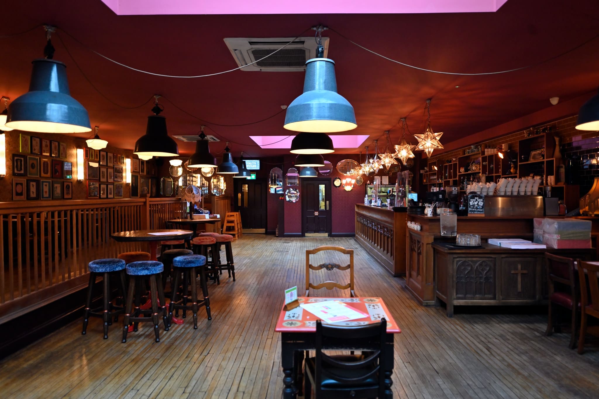 A cozy, warmly-lit pub called Conto features wooden floors and furniture, dark red walls, hanging lamps, star-shaped lights, framed photos, and a long bar counter lined with stools on the right side.