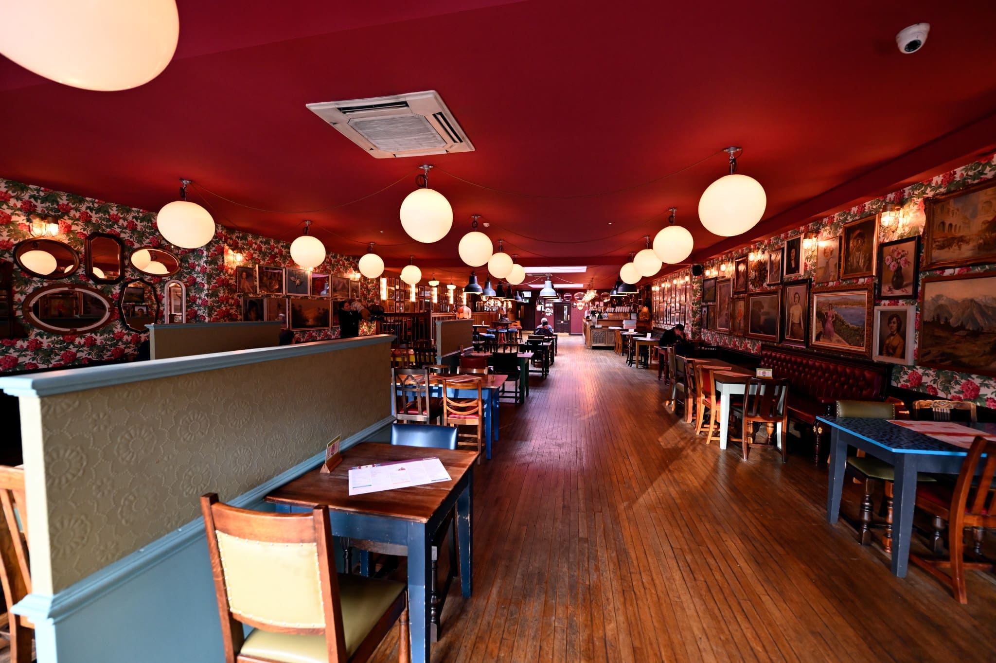 A cozy, vintage-style restaurant with wooden floors, floral wallpaper, round hanging lights, and eclectic wall art offers a classic Conto vibe. Wooden tables and chairs line the empty space, with a long aisle down the center.