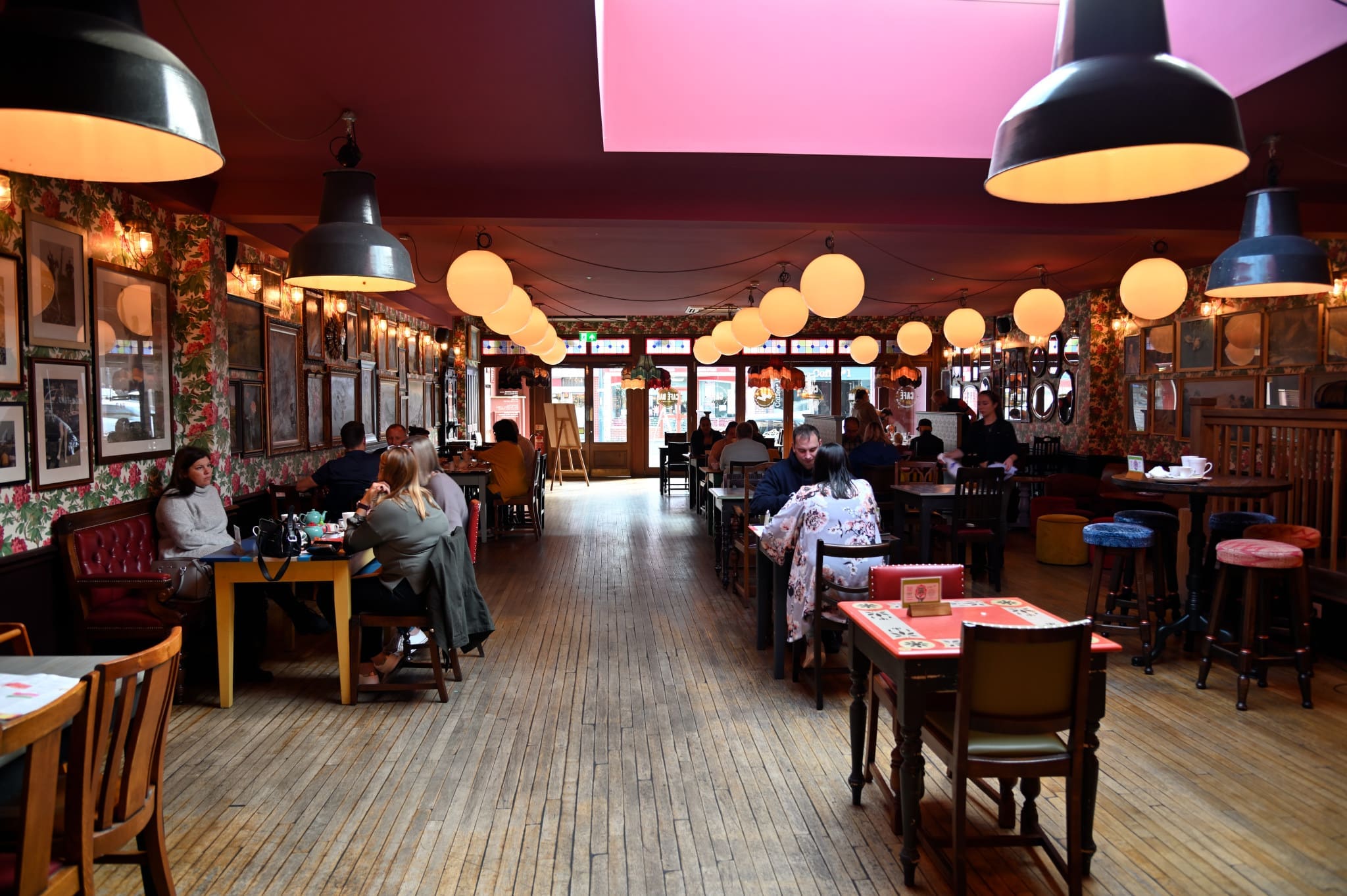 A cozy Conto restaurant with wooden floors, floral wallpaper, round hanging lights, and people dining at tables. The space is warmly lit, with framed pictures on the walls and a mix of empty and occupied tables.