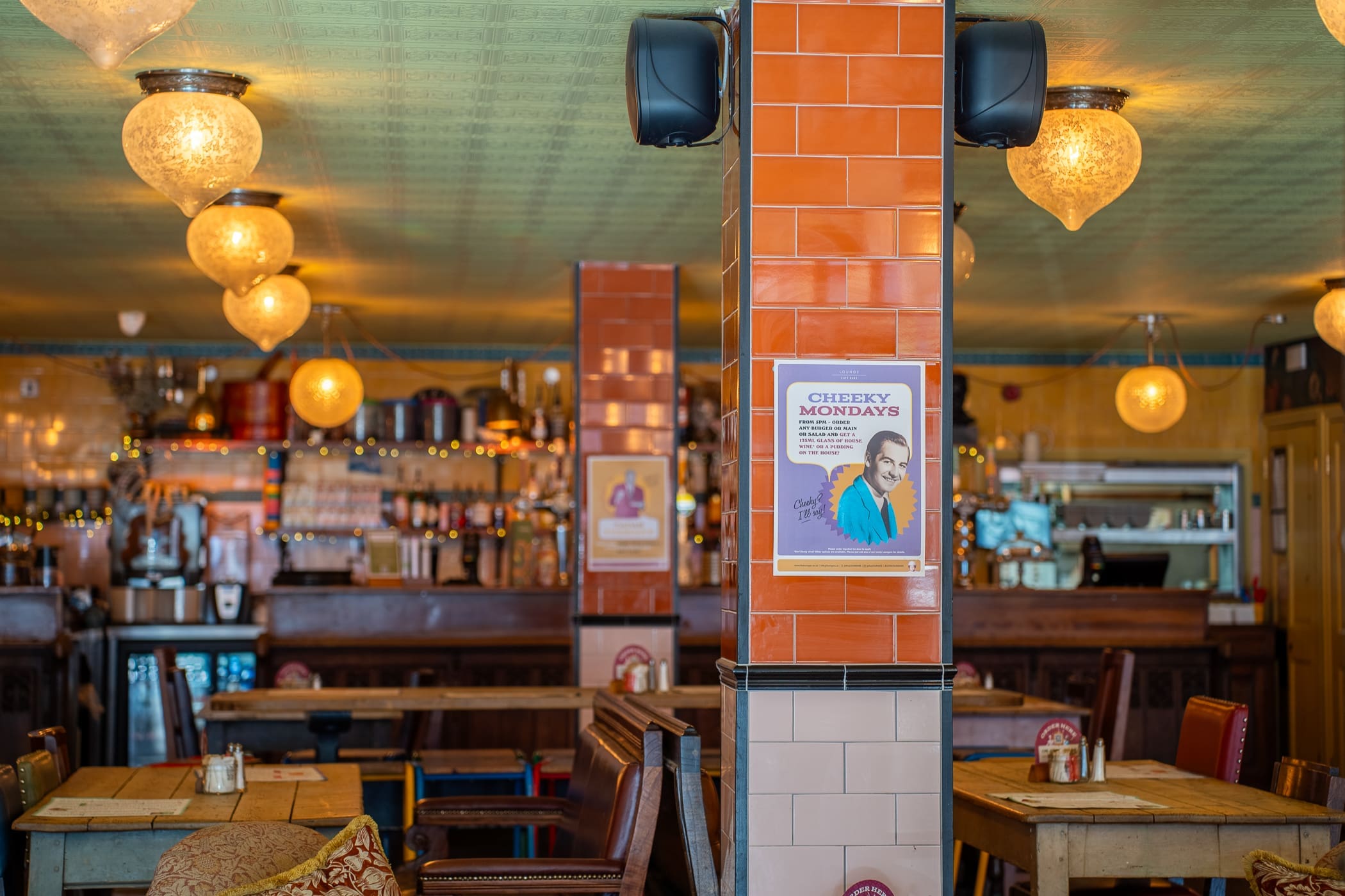 Cozy bar interior with wooden tables and benches, warm hanging lights, and a tiled column displaying a “Cheesy Mondays” poster featuring a Loco man. The bar counter and shelves with bottles are visible in the background.