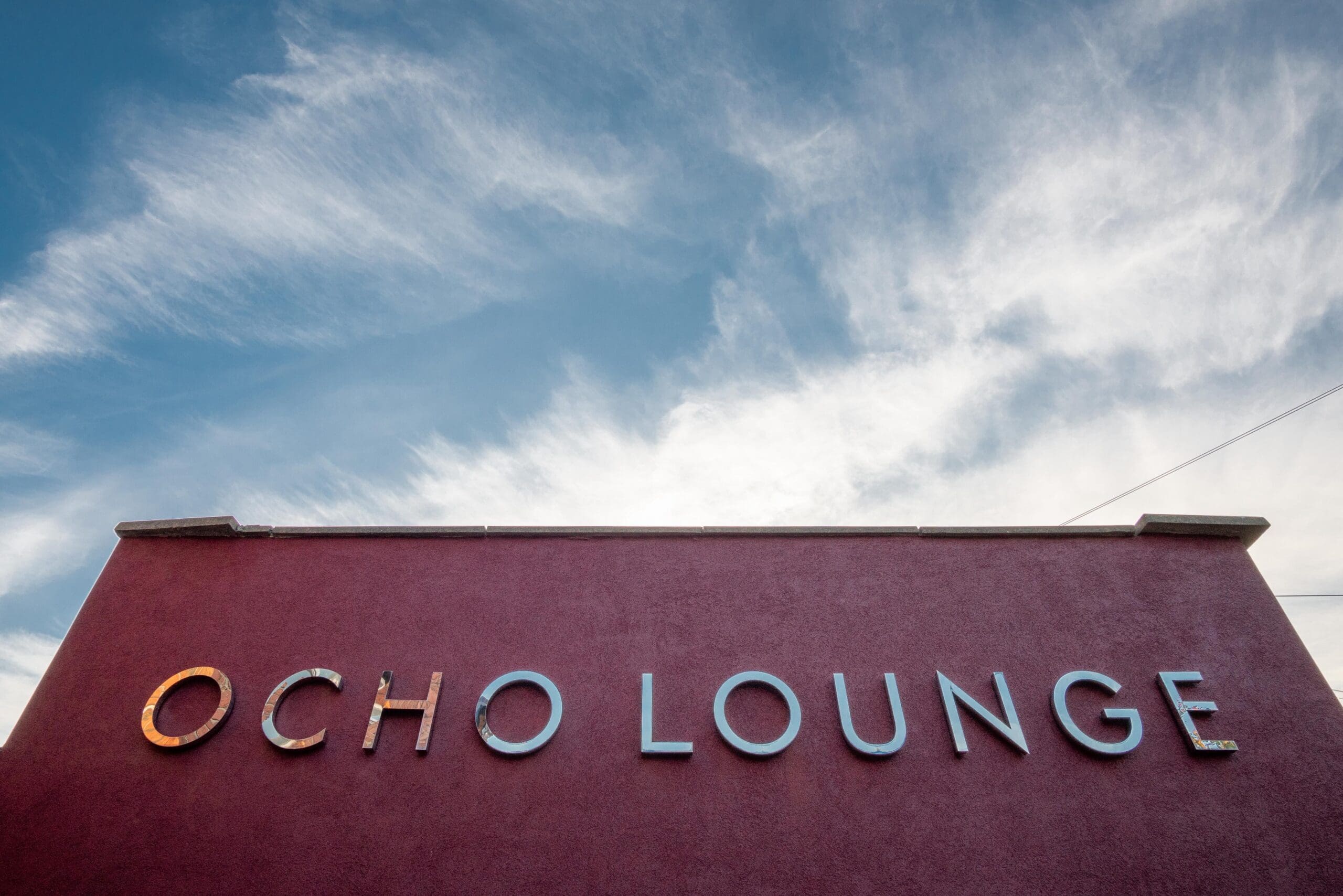 A red building named Ocho stands out, with metallic letters spelling "OCHO LOUNGE" against a blue sky brushed with wispy white clouds.