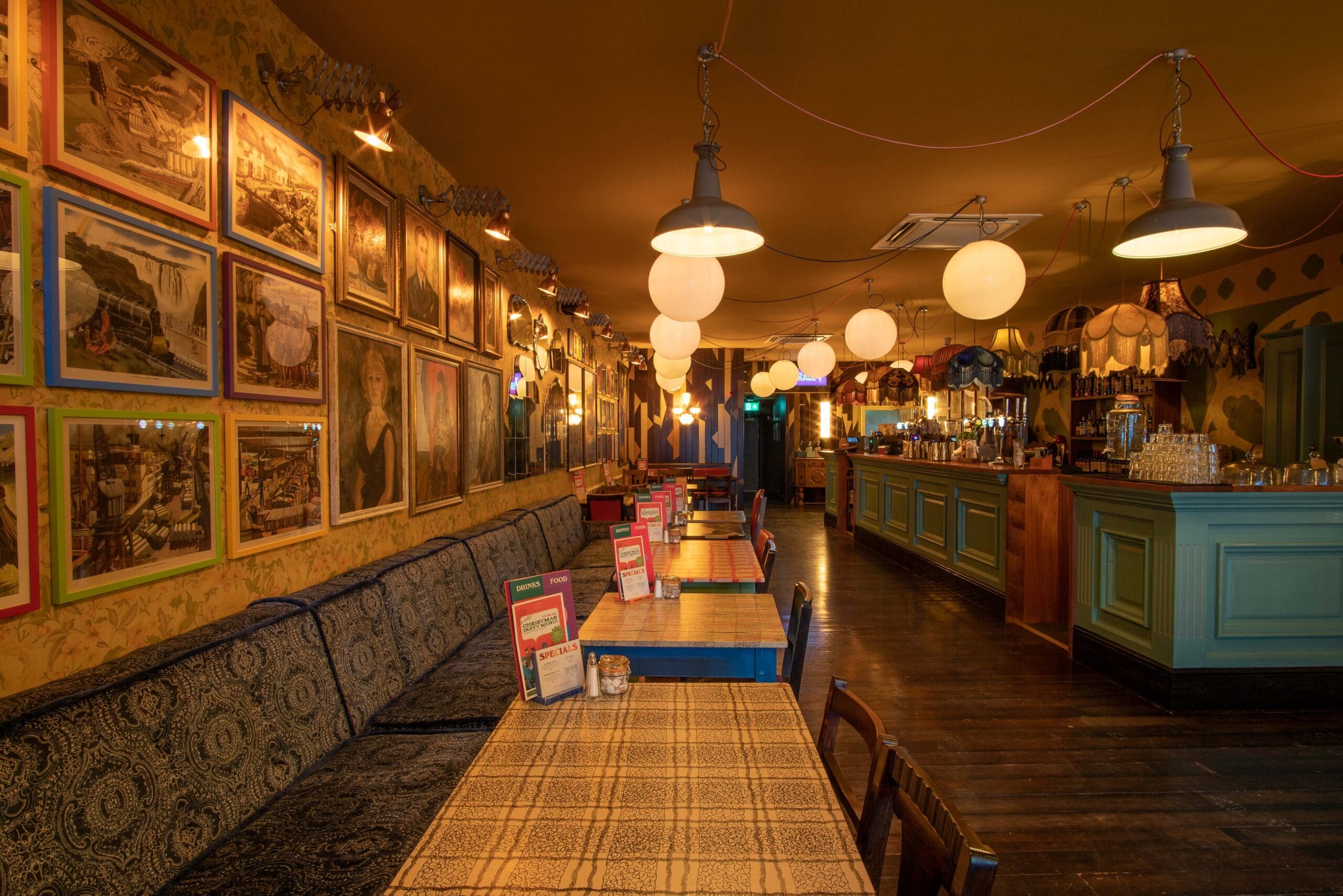 A cozy, warmly lit bar—Ocho—features framed pictures covering one wall, long cushioned bench seating, wooden tables with checkered cloths, and a blue-green bar counter in the background. Round pendant lights hang from the ceiling.
