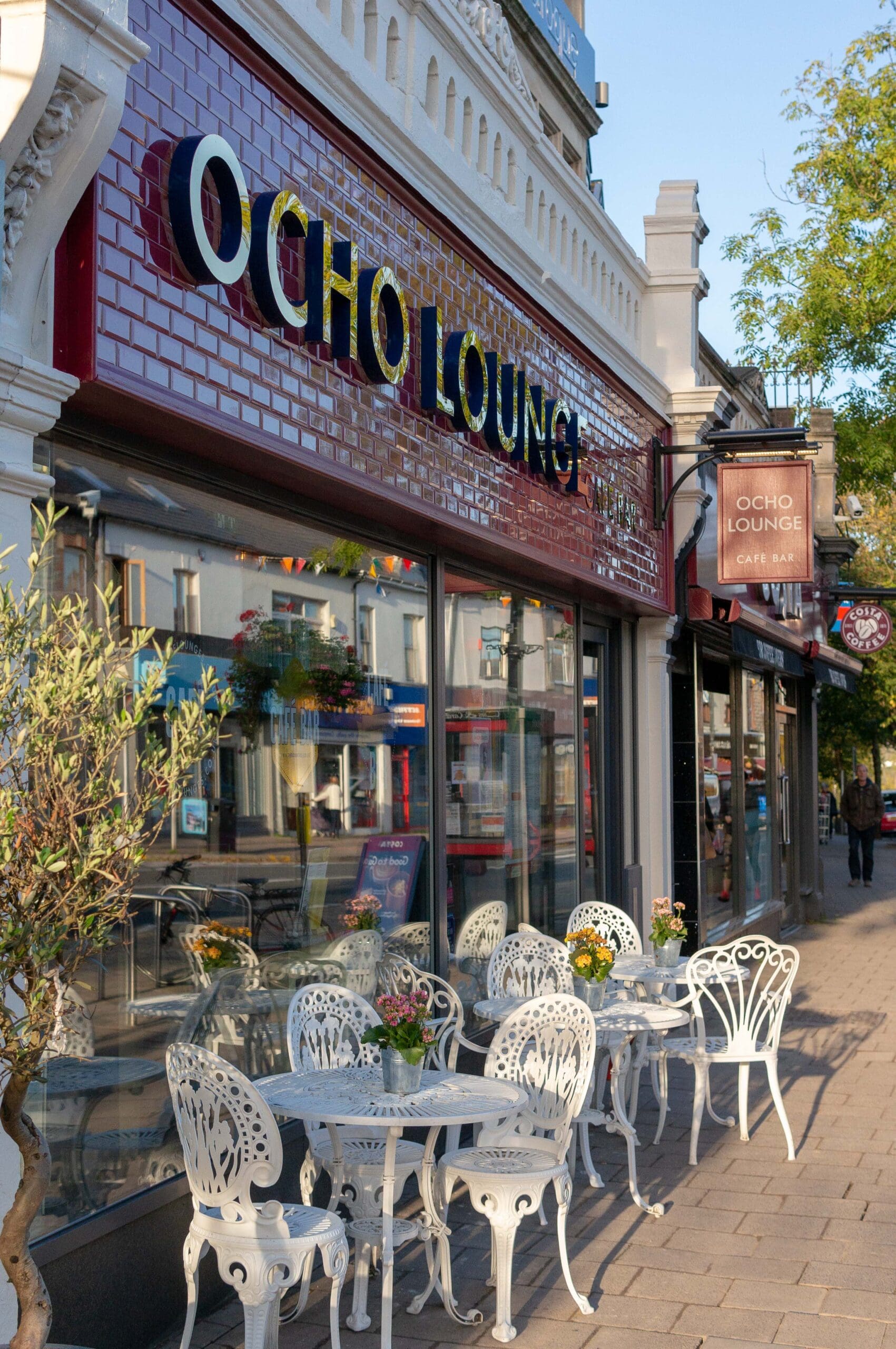 A sidewalk café called Ocho features ornate white metal tables and chairs outside a red tile façade. Flowers in vases top the tables, while trees and shops line the sunny street near the inviting Ocho Lounge sign.