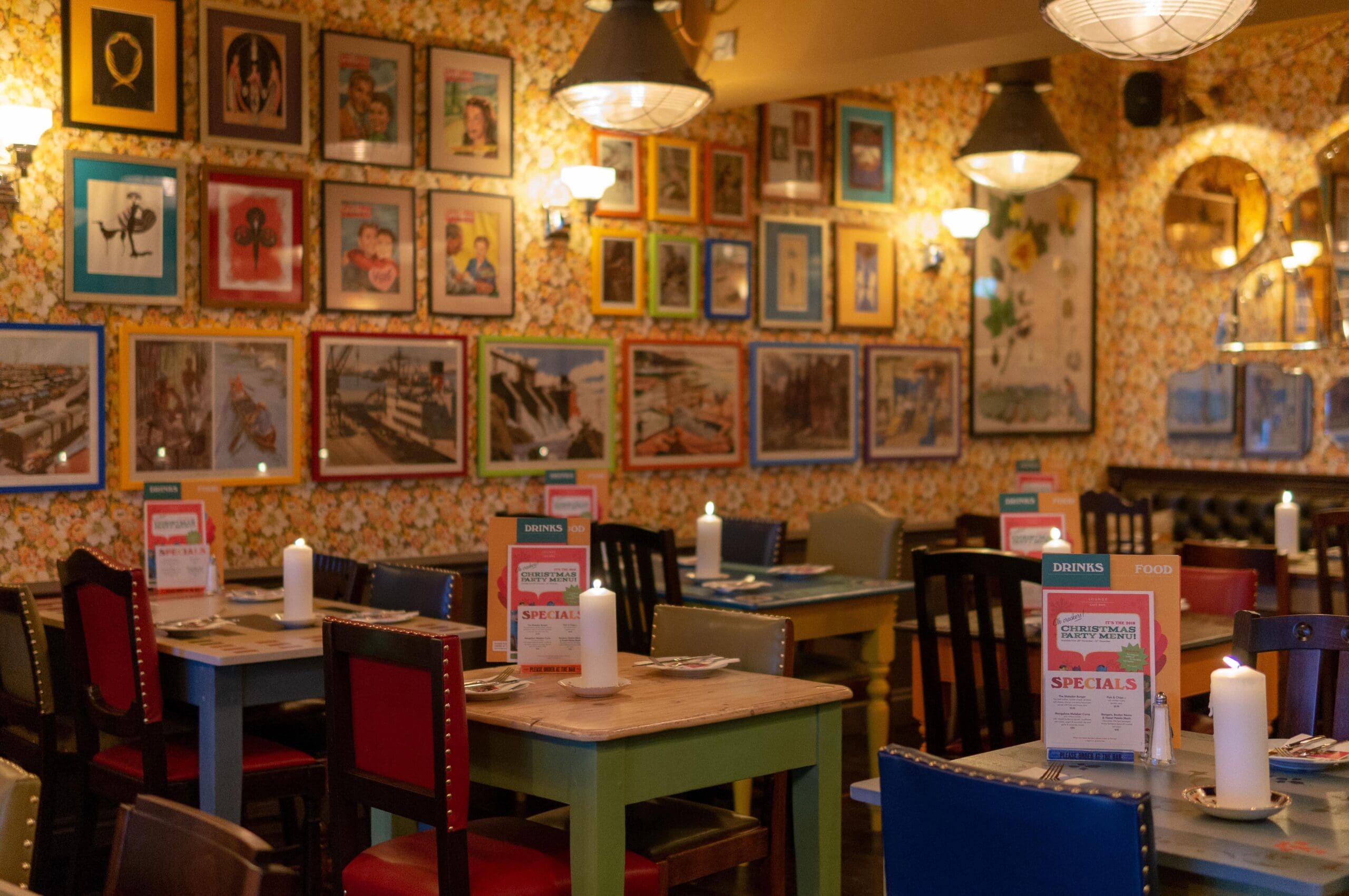 A cozy Ocho restaurant interior with colorful tables and chairs, lit candles on each table, and walls decorated with many framed pictures and vintage wallpaper. The atmosphere is warm and inviting.