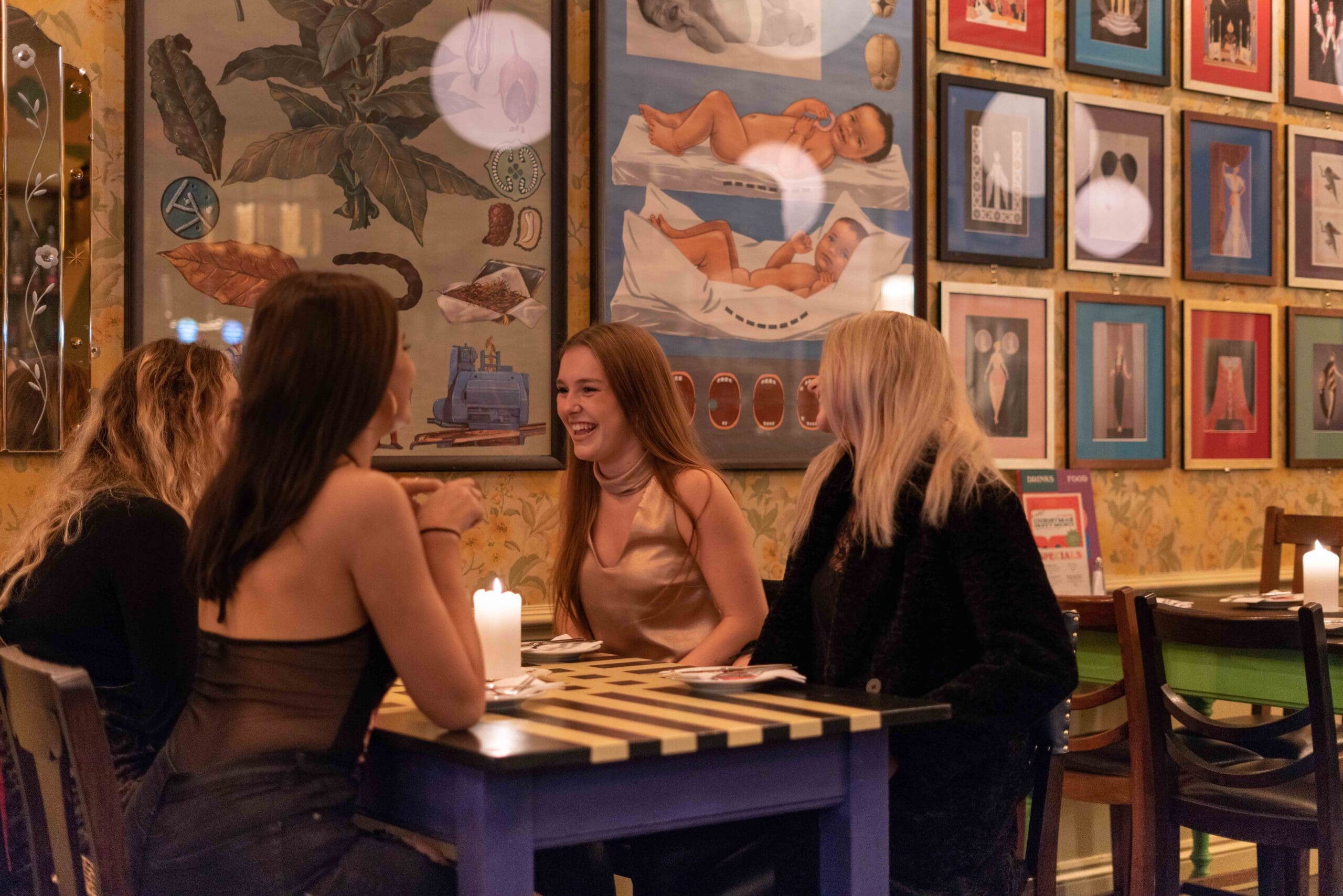 Four women sit around a table with lit candles in Ocho, a warmly lit restaurant. They chat and smile as colorful, eclectic framed artwork and photos decorate the walls.