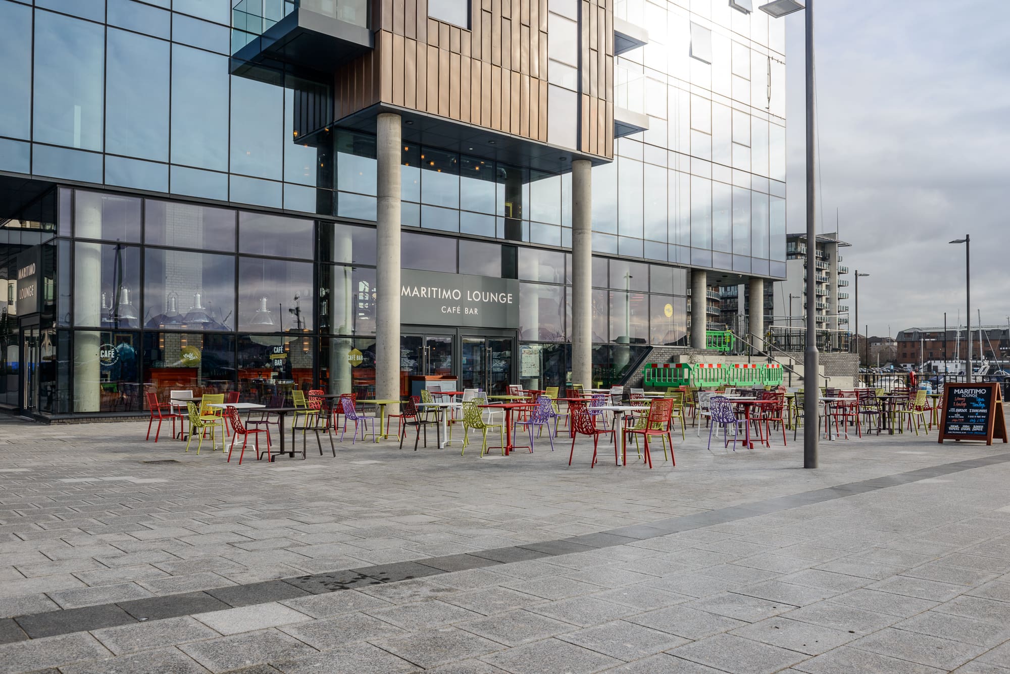 Outdoor seating area with colorful chairs and tables in front of Maritimo Lounge Café Bar, a modern glass-fronted Maritimo building, on a paved plaza under a cloudy sky.