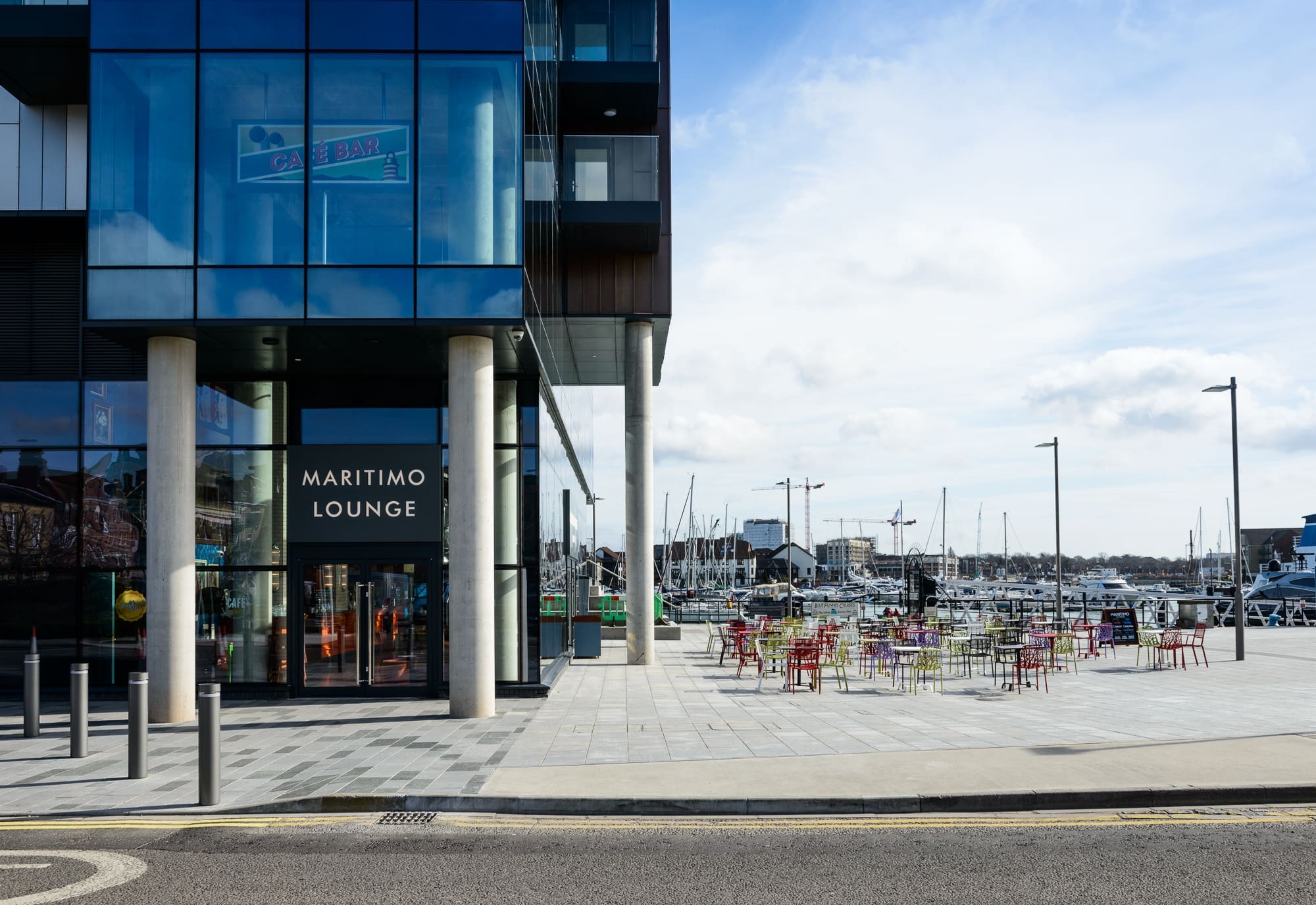 Glass-fronted building with a sign reading "Maritimo Lounge" beside an outdoor seating area with colorful chairs and tables. The Maritimo overlooks a marina dotted with boats and masts under a cloudy sky.
