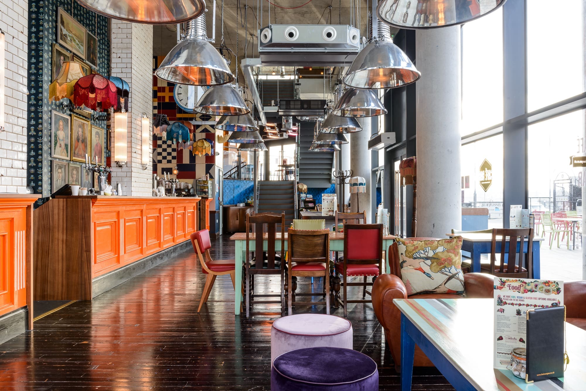 A colorful, eclectic café interior inspired by Maritimo, with mismatched chairs, a bright orange bar, modern light fixtures, and playful décor like patterned pillows and hanging lampshades. Natural light fills the vibrant space.