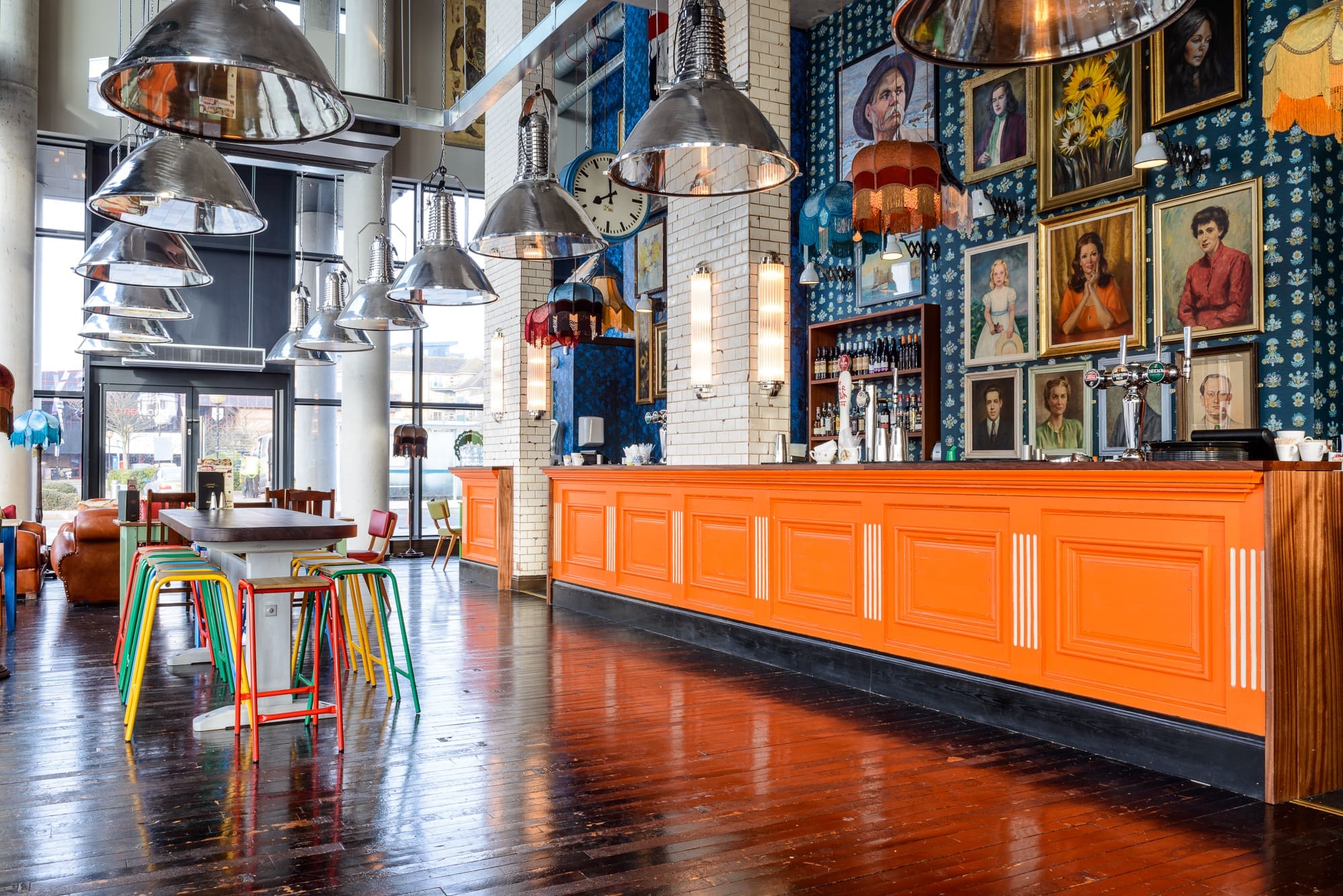A brightly lit Maritimo bar with an orange counter, colorful stools, pendant lights, and a wall adorned with eclectic vintage portraits, wallpaper, and lampshades. Large windows let sunlight spill onto the glossy wooden floor.