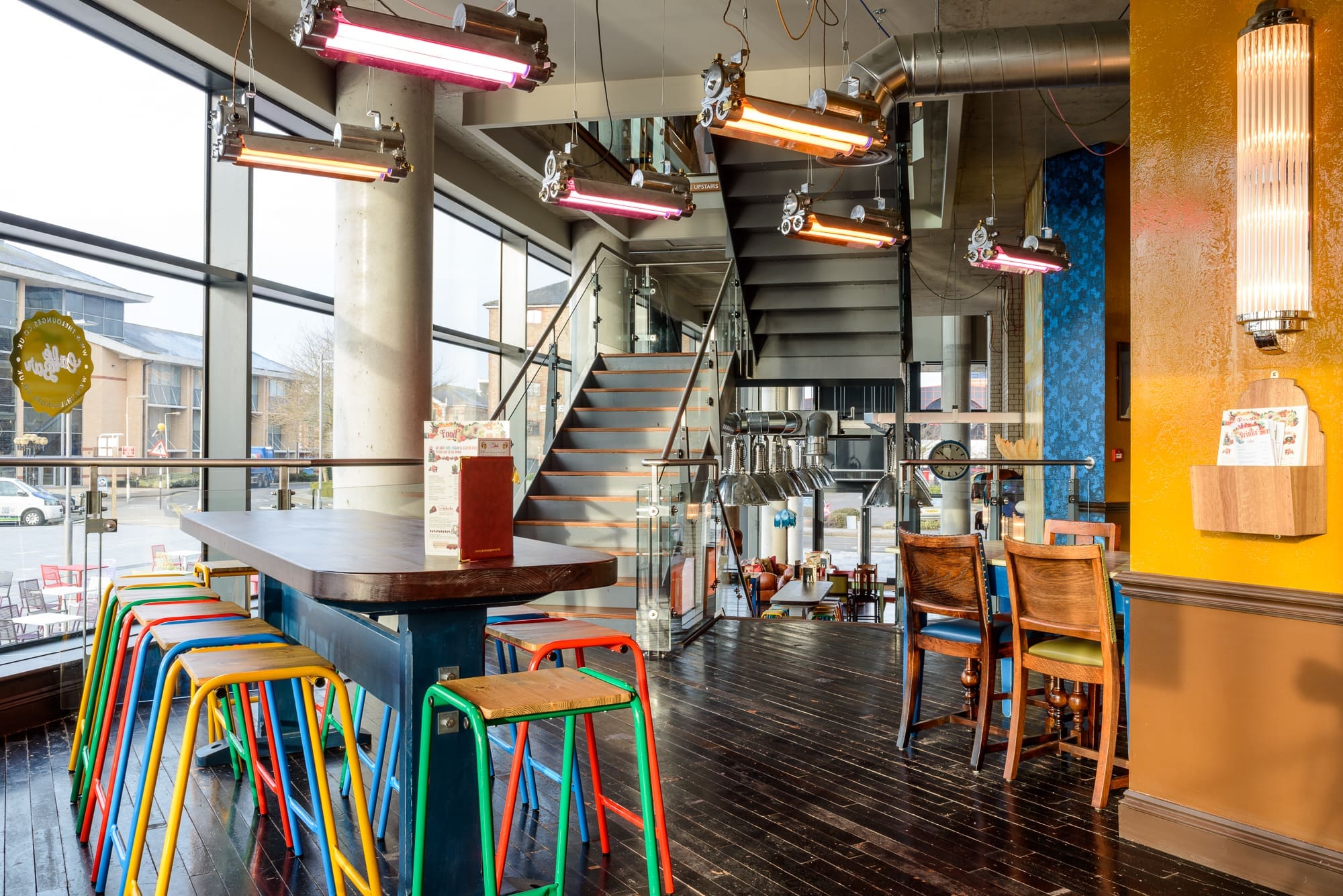 A modern café interior with colorful Maritimo metal stools around a tall table, wooden flooring, large windows, and a staircase to an upper level. Bright pendant lights hang from the ceiling, creating a vibrant atmosphere.