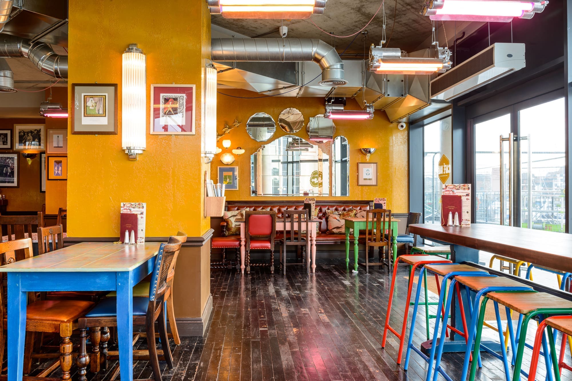 A brightly colored café interior with yellow walls, eclectic framed art, round mirrors, colorful tables and chairs, and large windows letting in natural light. Maritimo-inspired bar stools line a high wooden counter on the right.