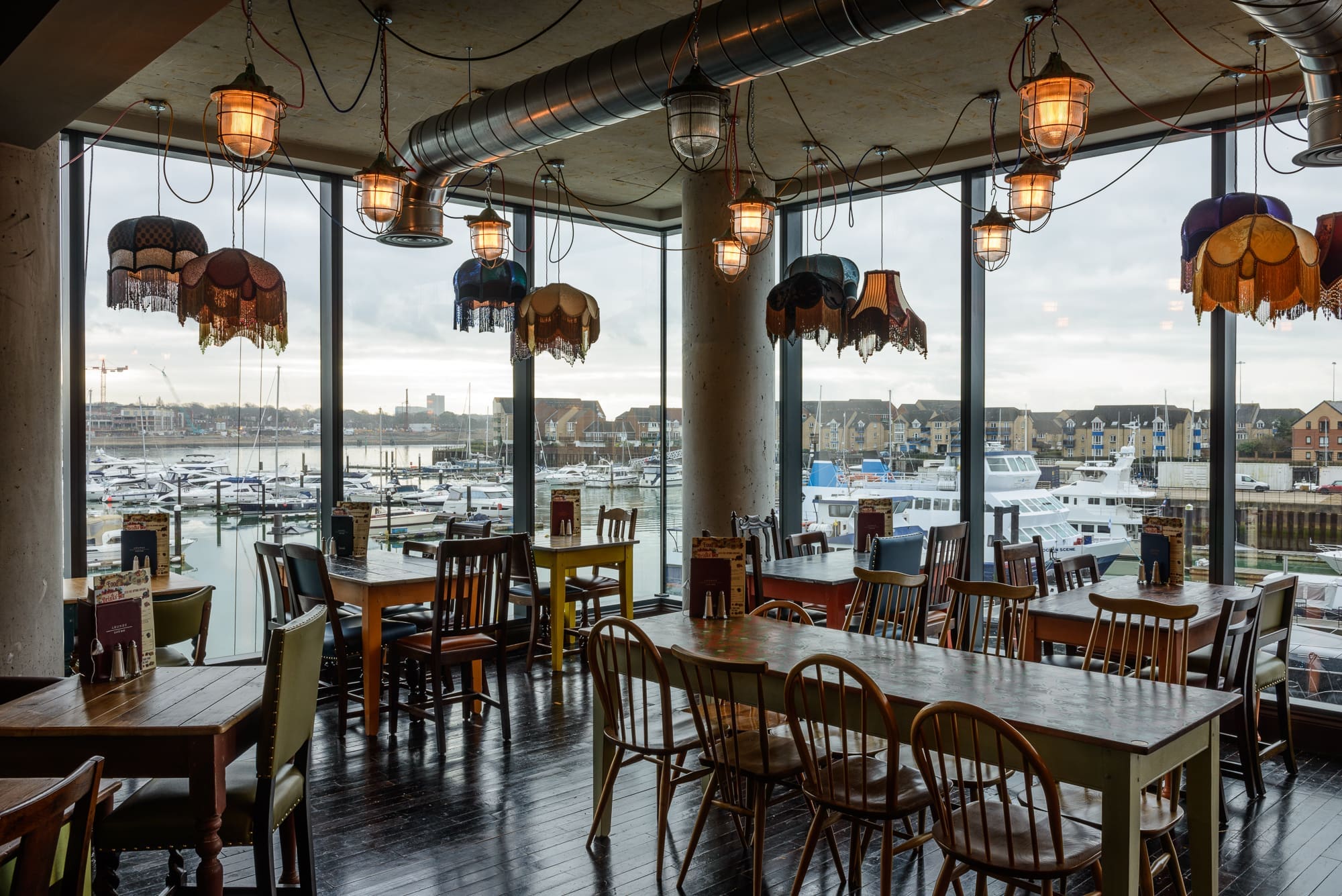 A cozy Maritimo restaurant with vintage lamps, wooden tables, and mismatched chairs overlooks a marina filled with boats, seen through large floor-to-ceiling windows on a cloudy day.