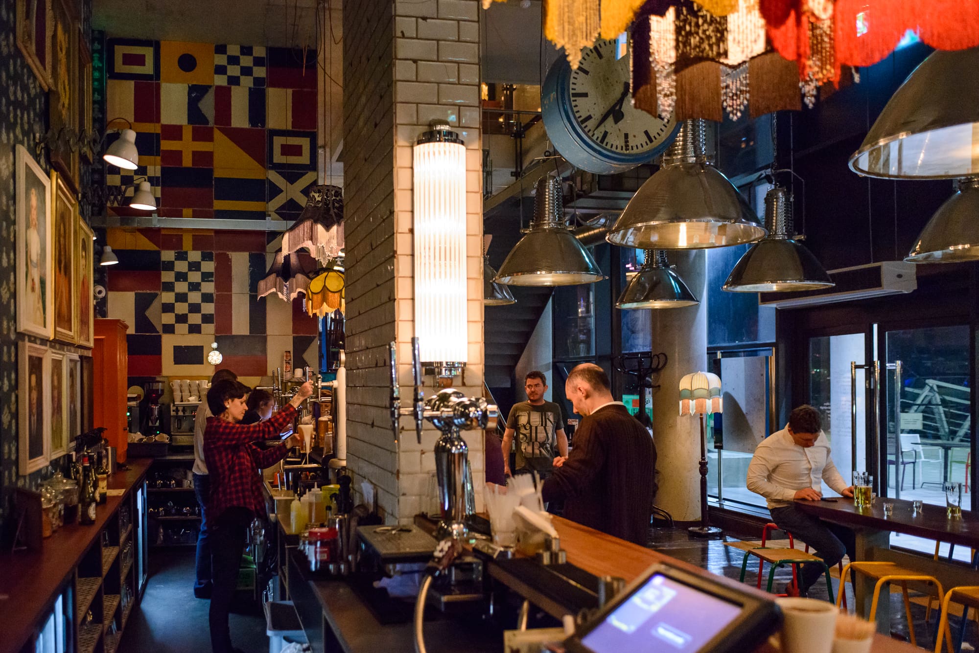 A cozy Maritimo bar with colorful wall art and industrial lighting. A bartender prepares drinks behind the counter while a few patrons sit at the bar and tables, some talking and others using their phones.