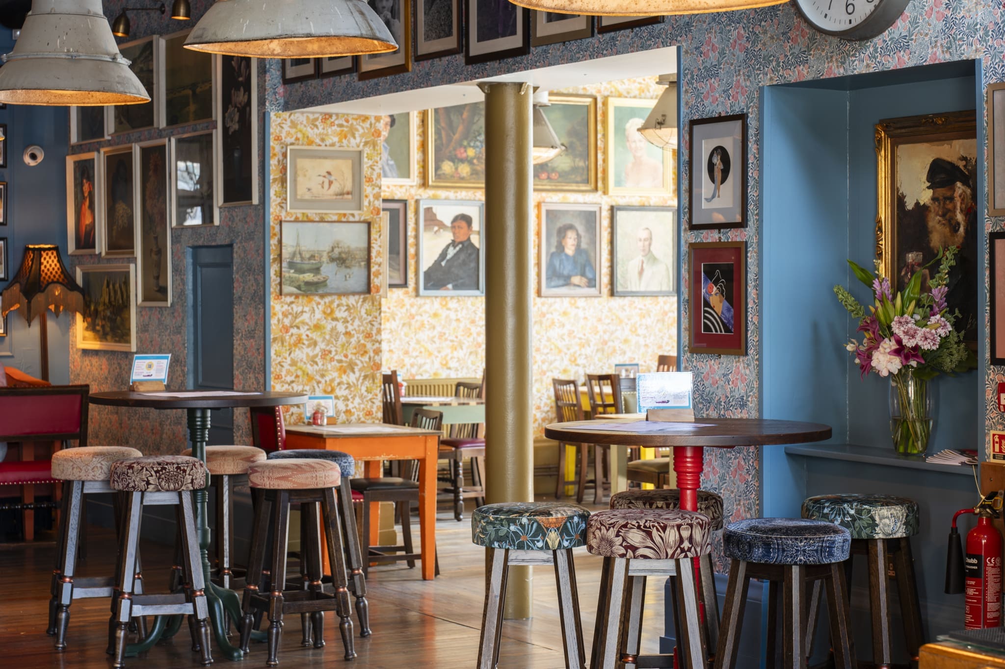 A cozy, eclectic cafe interior reminiscent of a Palacio, with patterned wallpaper, round wooden tables, mismatched upholstered stools, and walls adorned with framed portraits and artwork. A bouquet of flowers sits on a shelf.