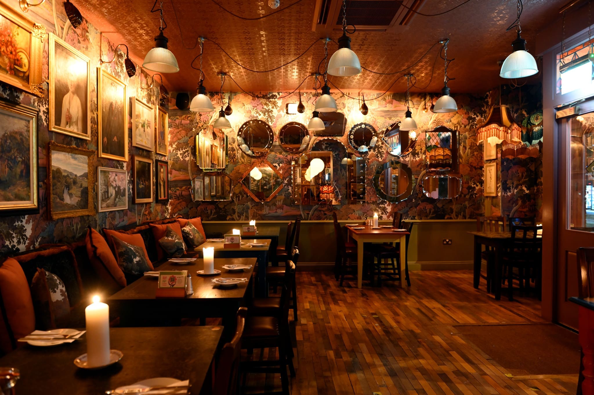 A cozy, dimly lit restaurant in Porto with wooden floors, tables set for dining, candles on each table, eclectic framed art and mirrors on patterned walls, and warm ambient lighting from pendant lamps overhead.