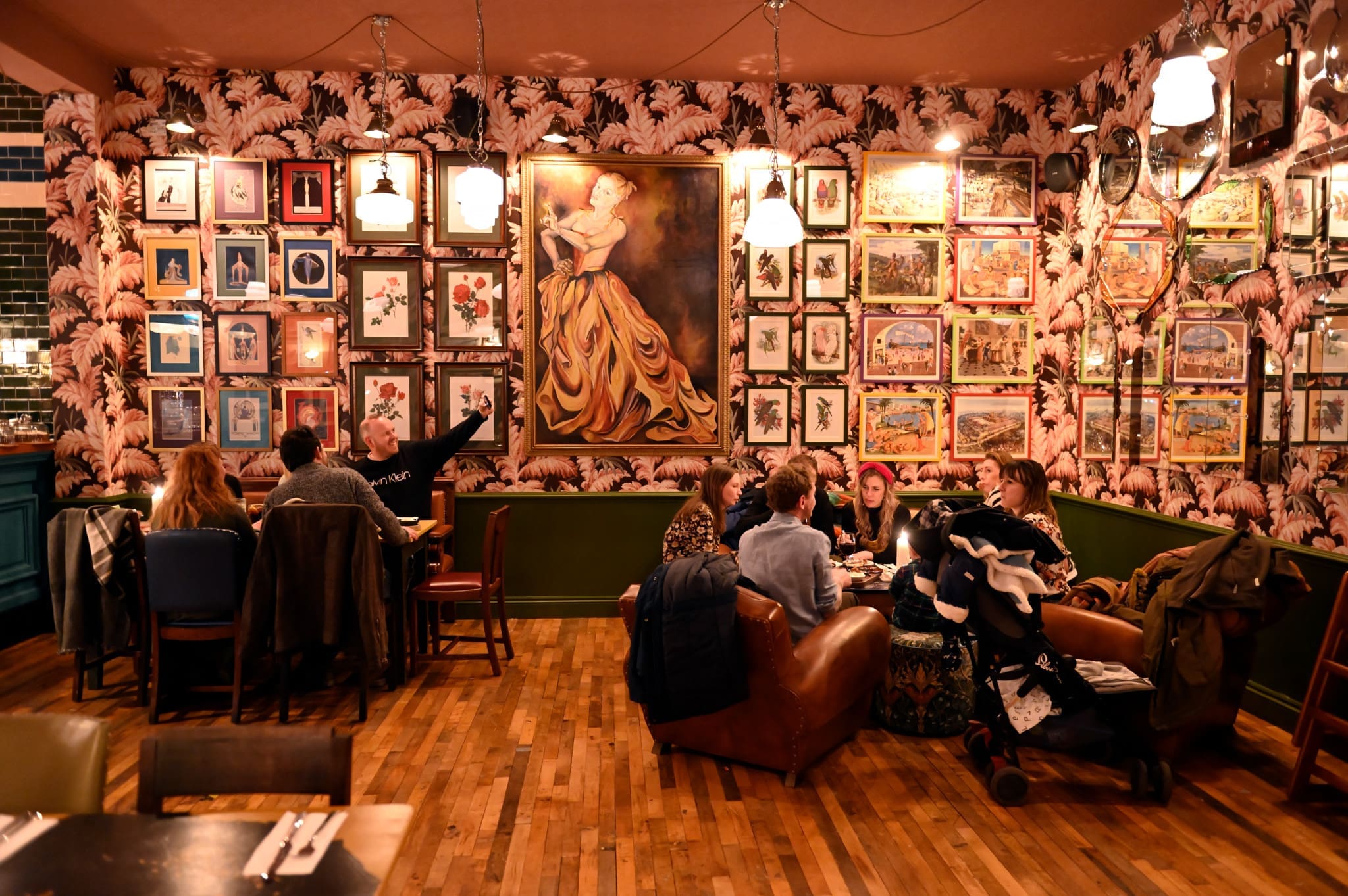 People sit at tables and chat in a cozy Porto restaurant with wooden floors and eclectic decor. The walls are covered with framed artwork, including a large painting of a woman in a yellow dress at the center.