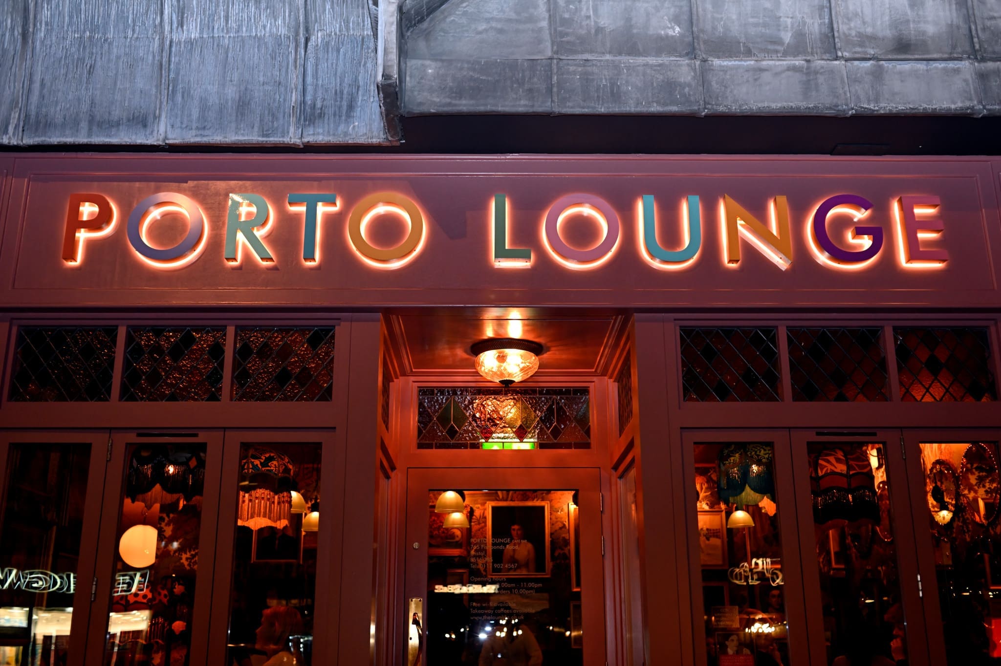 A neon sign reading "PORTO LOUNGE" glows above the entrance of a cozy, warmly lit Porto bar at night, with reflections shimmering in the glass doors and windows.