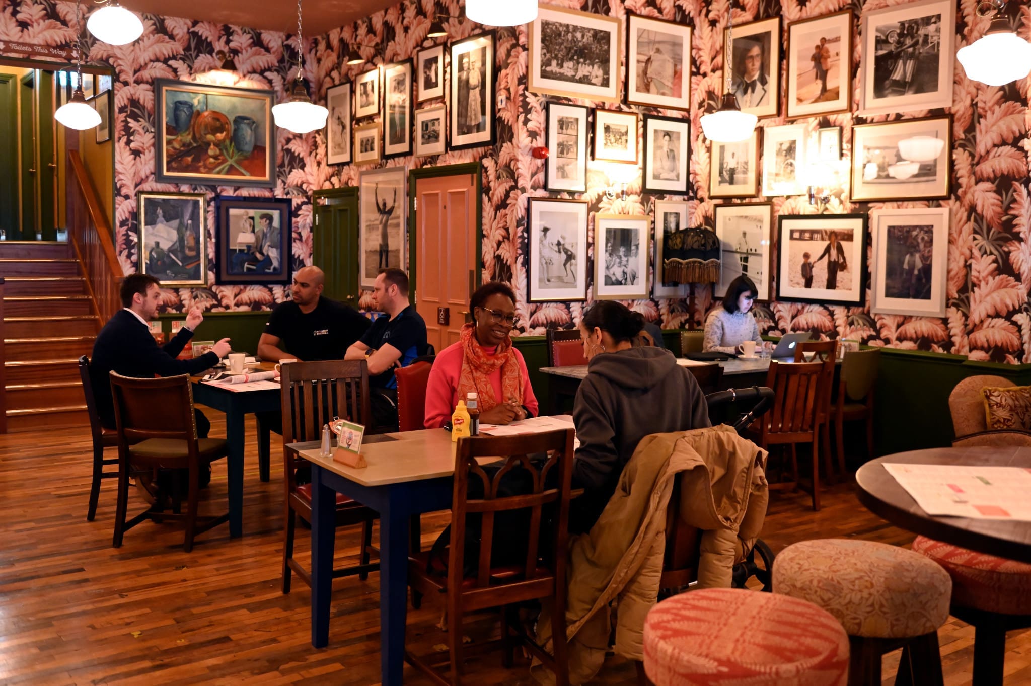 People sit at tables in a cozy Porto café with wooden floors, patterned wallpaper, and framed photos covering the walls. Some are talking, others use laptops. Warm lighting creates a relaxed atmosphere.
