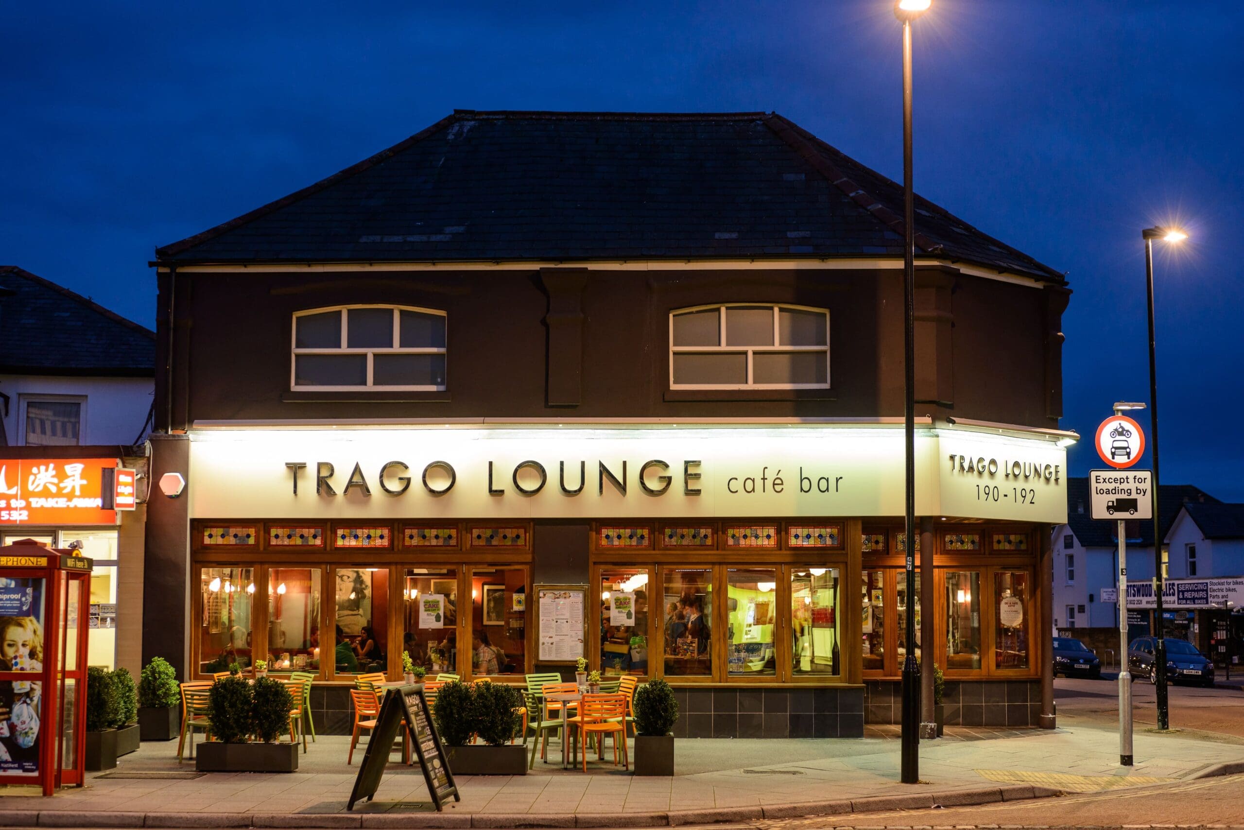 A warmly lit café bar called Trago Lounge sits on a street corner at dusk. Outdoor tables and chairs are arranged by the entrance, and people can be seen inside through large windows. Street lamps cast a cozy glow over Trago’s inviting scene.