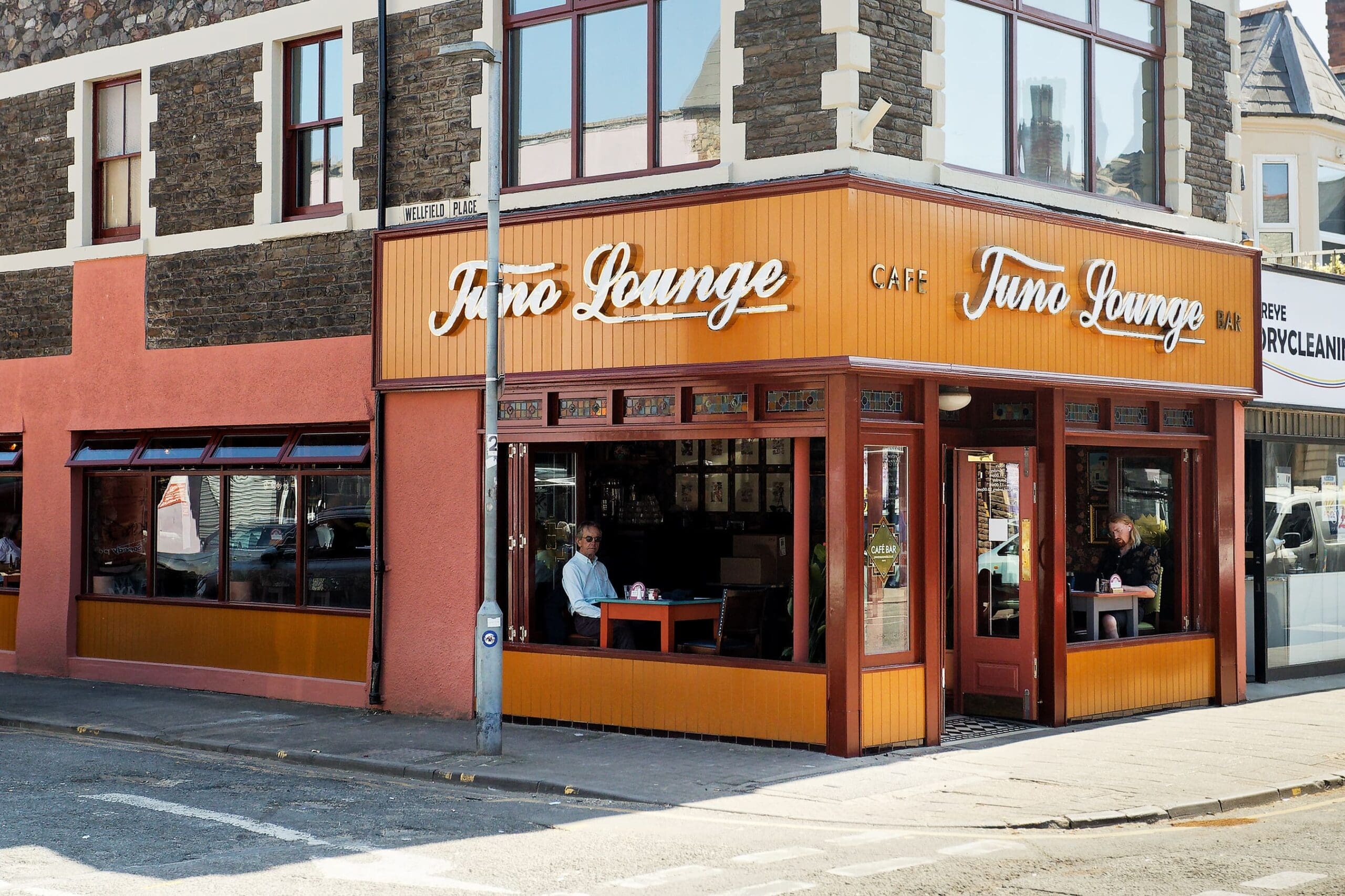Street view of Juno Lounge café with large windows, orange trim, and two people sitting inside near the front. The building sits on a quiet street with stone and red-painted exterior walls.
