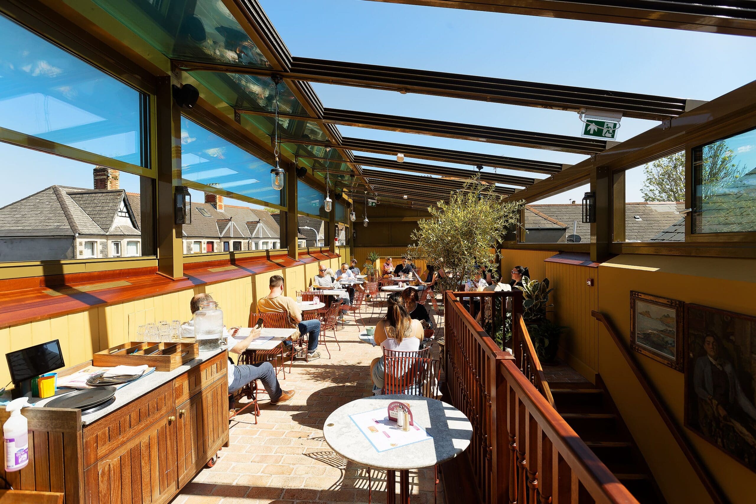 People dining at Juno’s rooftop restaurant with glass walls and a partially open roof on a sunny day. Some tables are empty, while others have groups chatting and eating. The bright, airy setting features potted plants throughout.